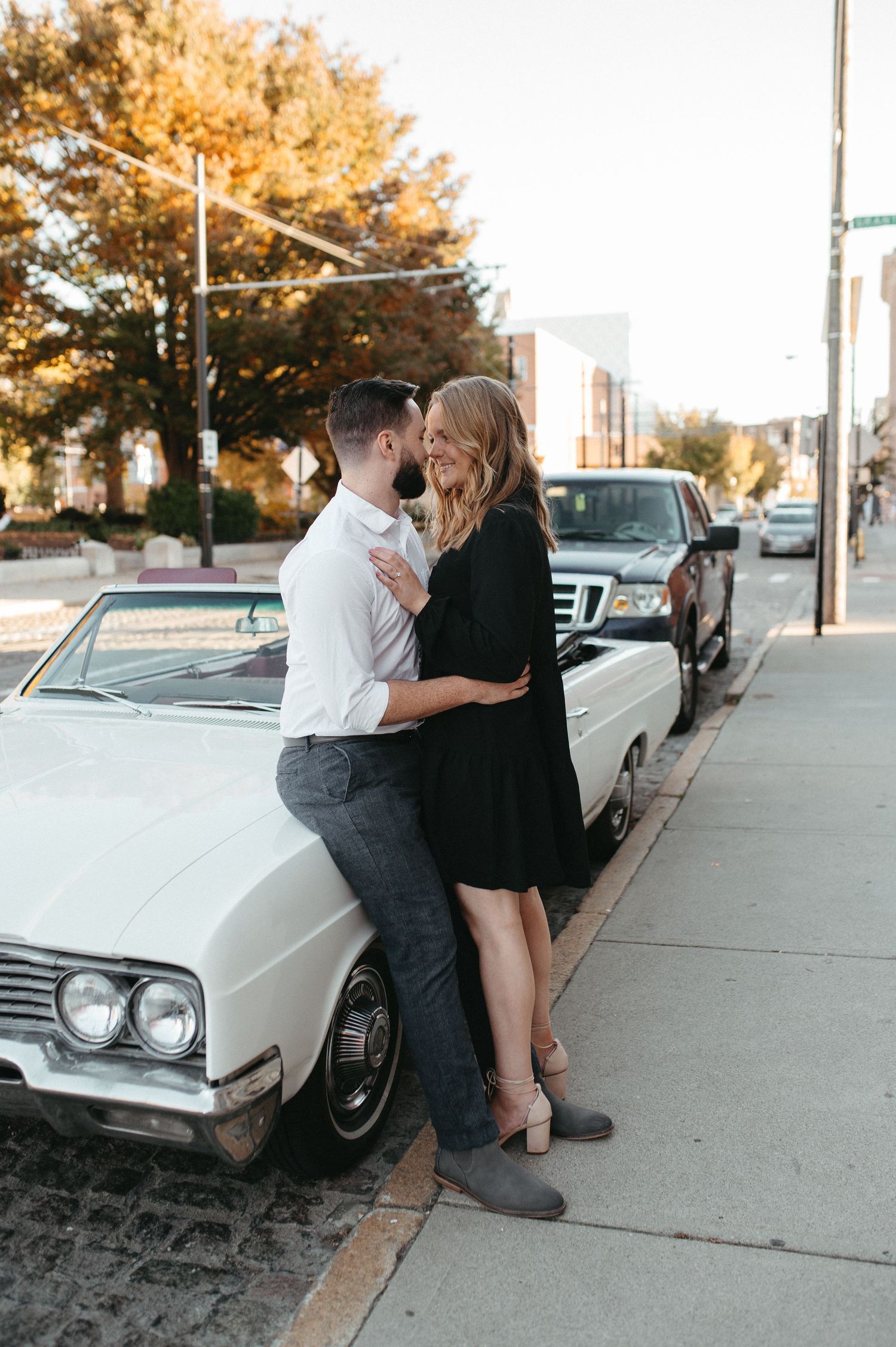 Couple shares an intimate moment leaning against a vintage white automobile on a city sidewalk.