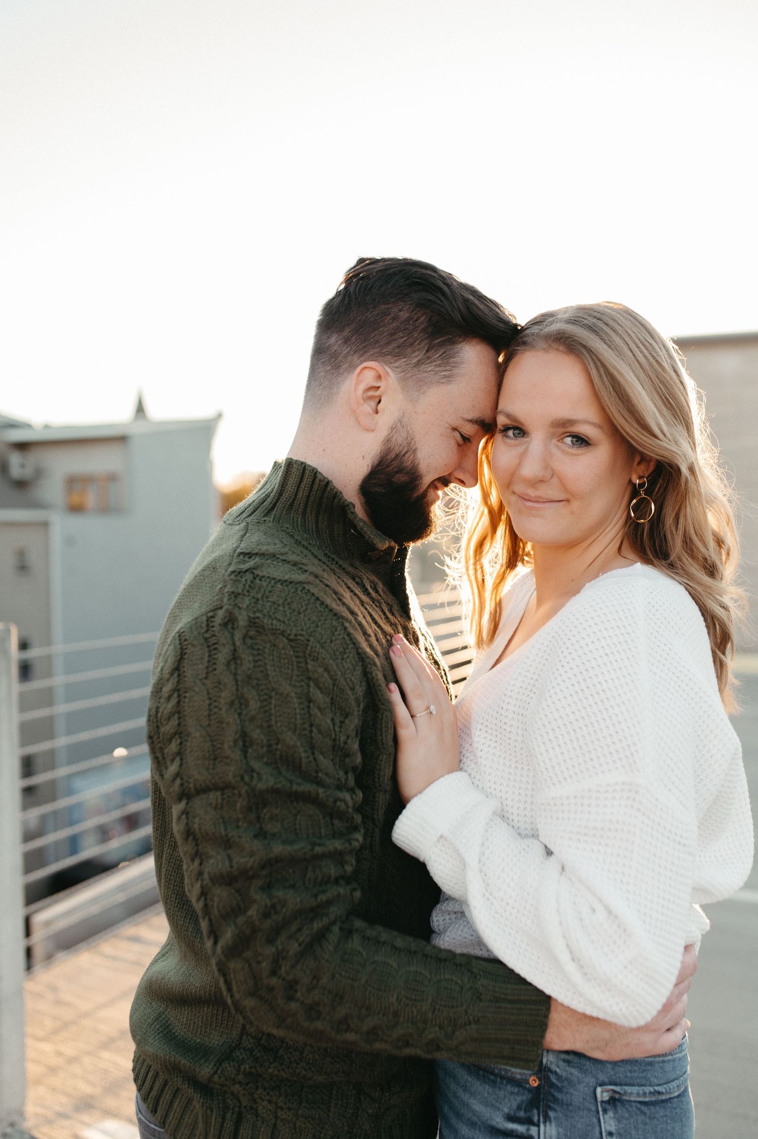 Couple embracing in warm sunlight wearing cozy sweaters during engagement photoshoot.