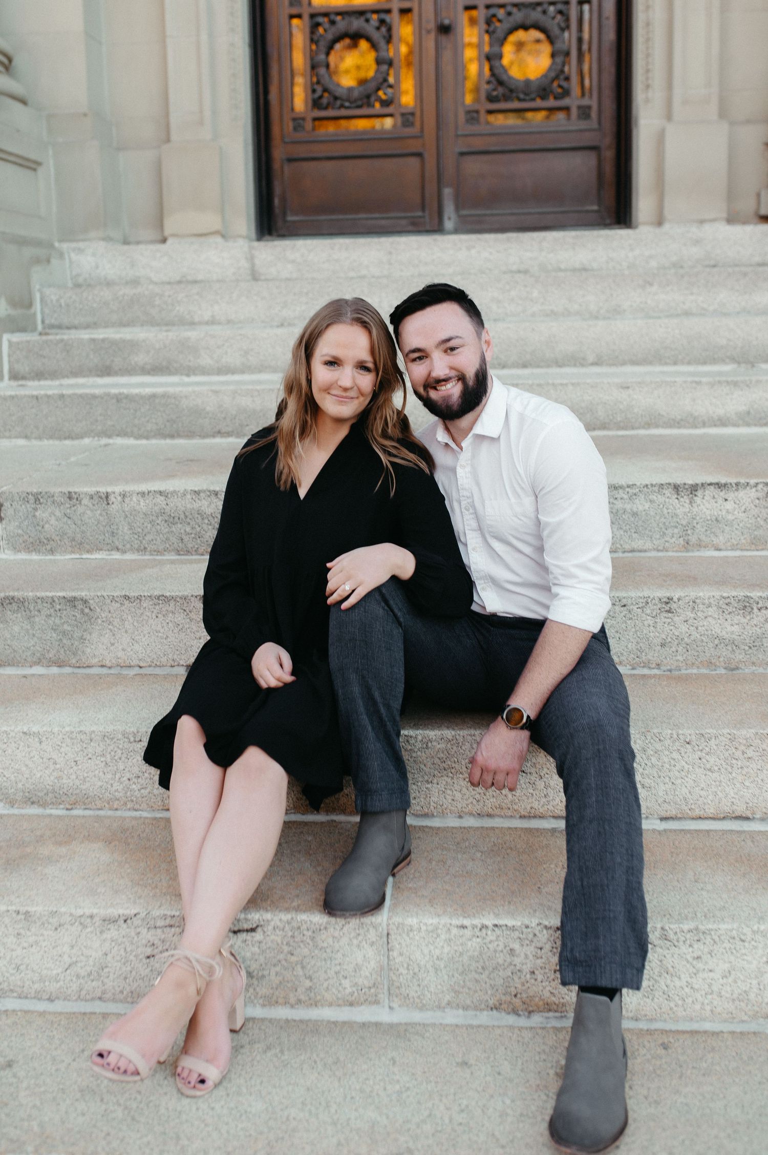 A couple sits together on stone steps in front of ornate wooden doors during an engagement photoshoot.