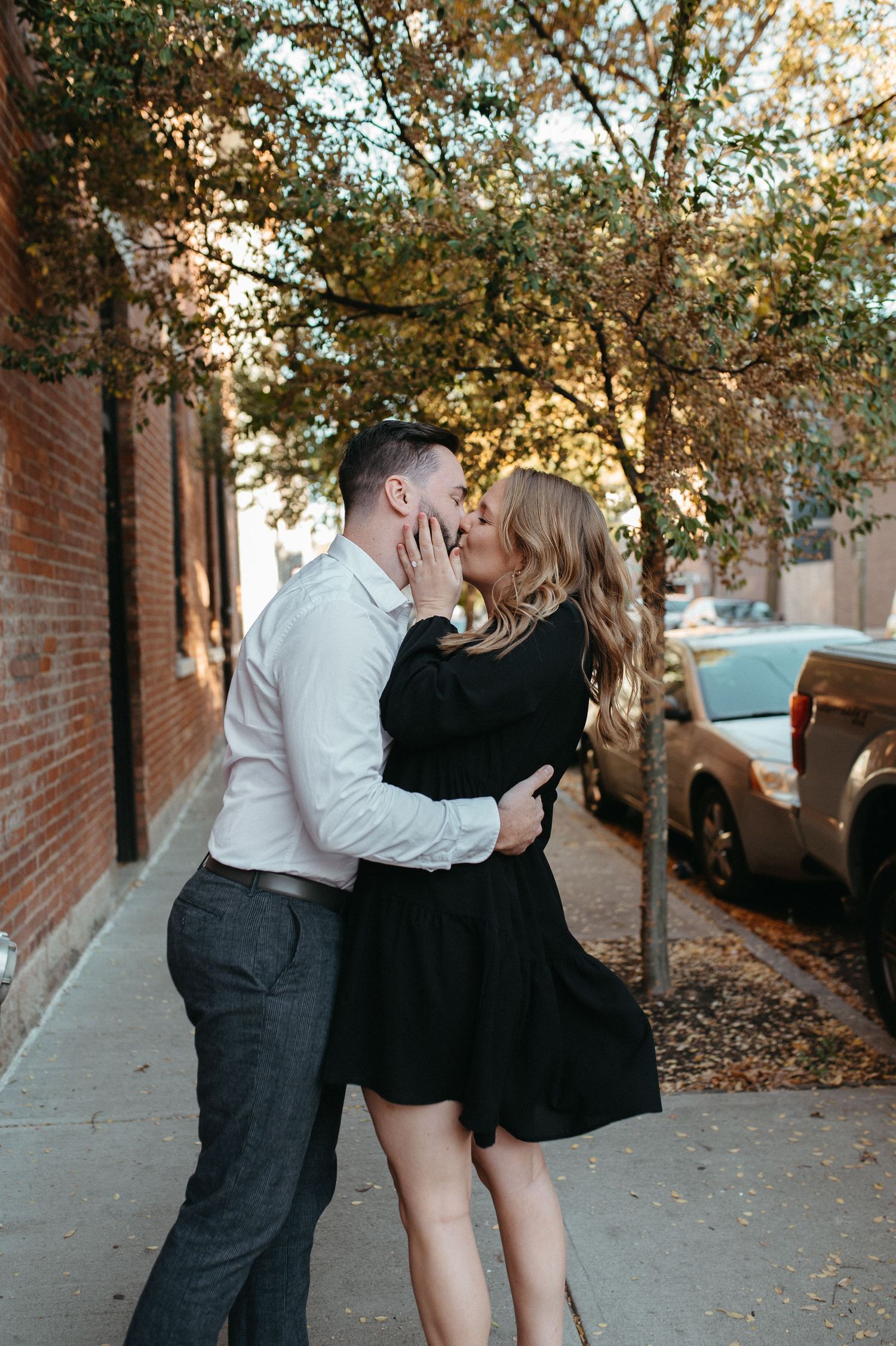 Couple sharing a romantic kiss on a sidewalk with autumn trees and parked cars in the background.