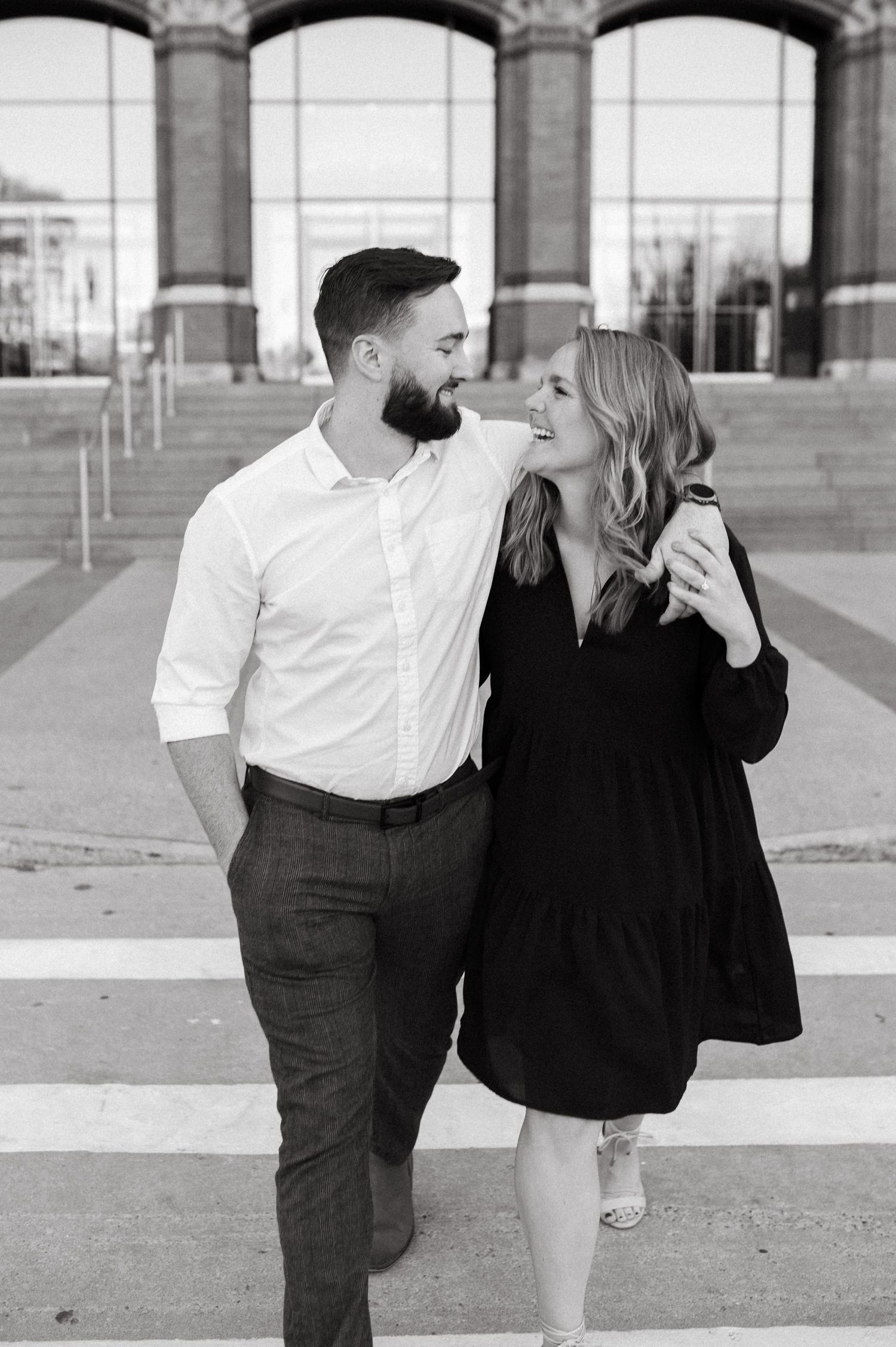 A black and white portrait of a couple laughing together in front of an architectural backdrop.