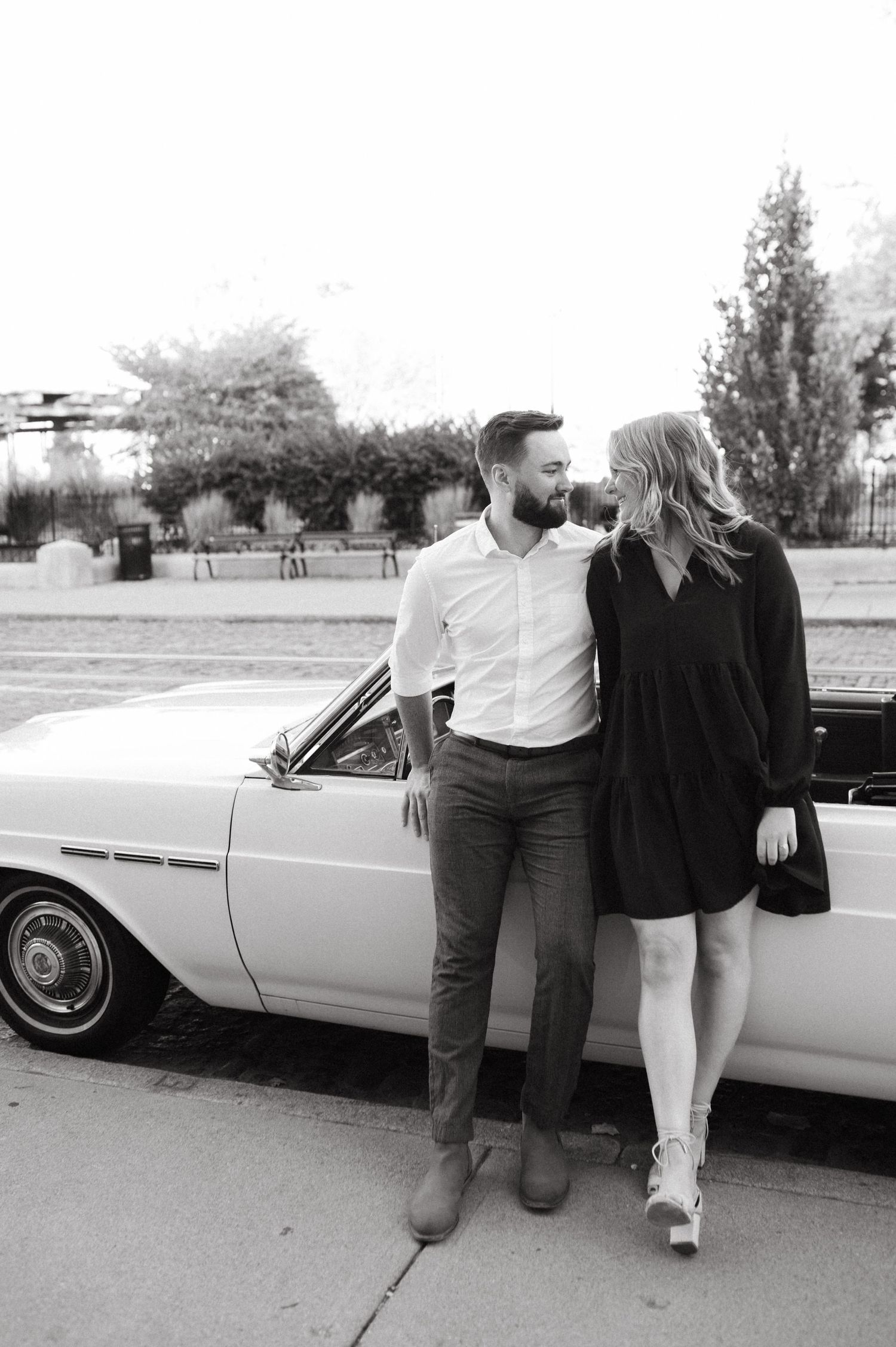 A black and white photo of a couple leaning against a vintage white car during a photoshoot.