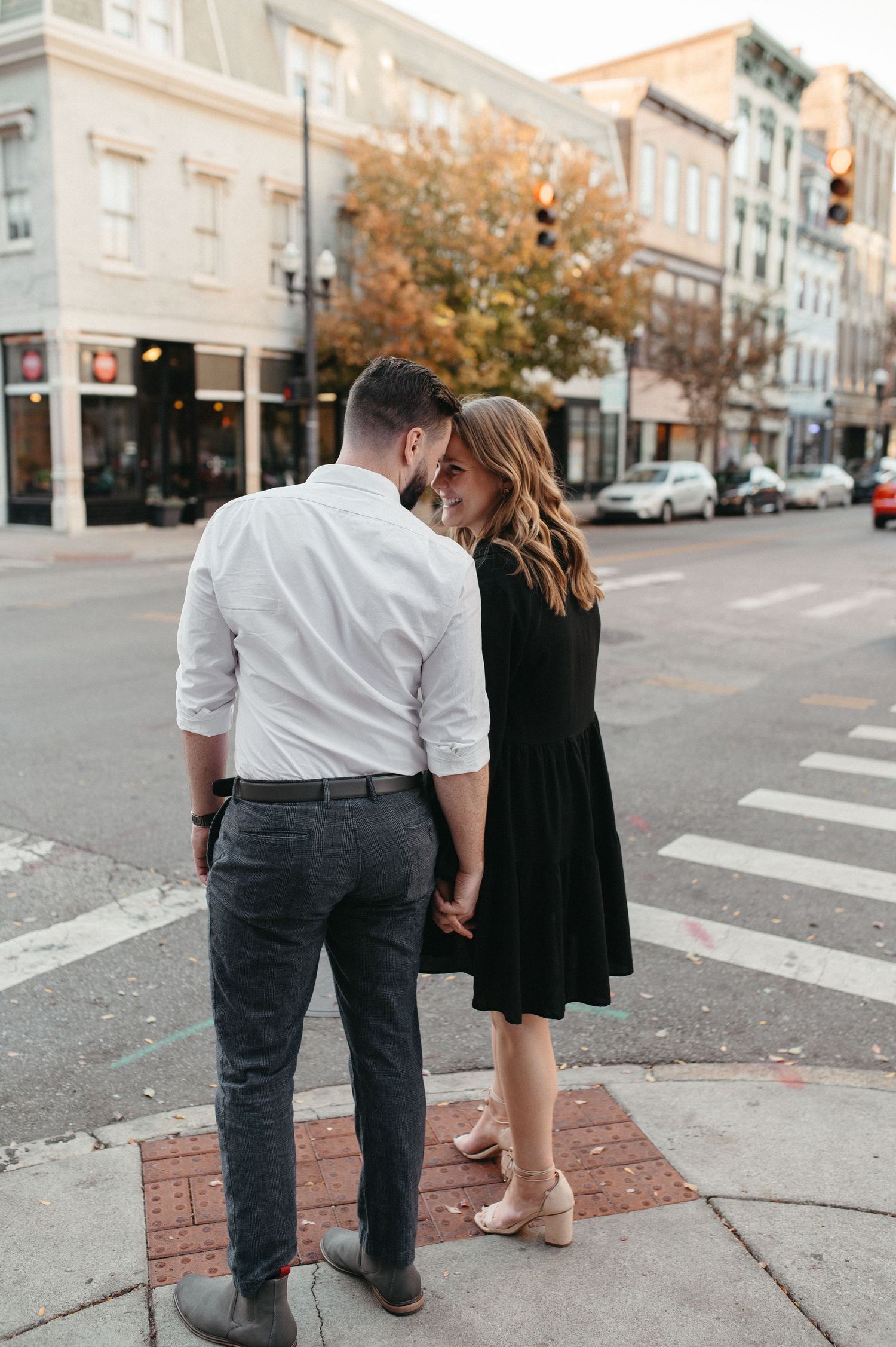 A couple embraces on a brick sidewalk corner in a historic downtown setting.