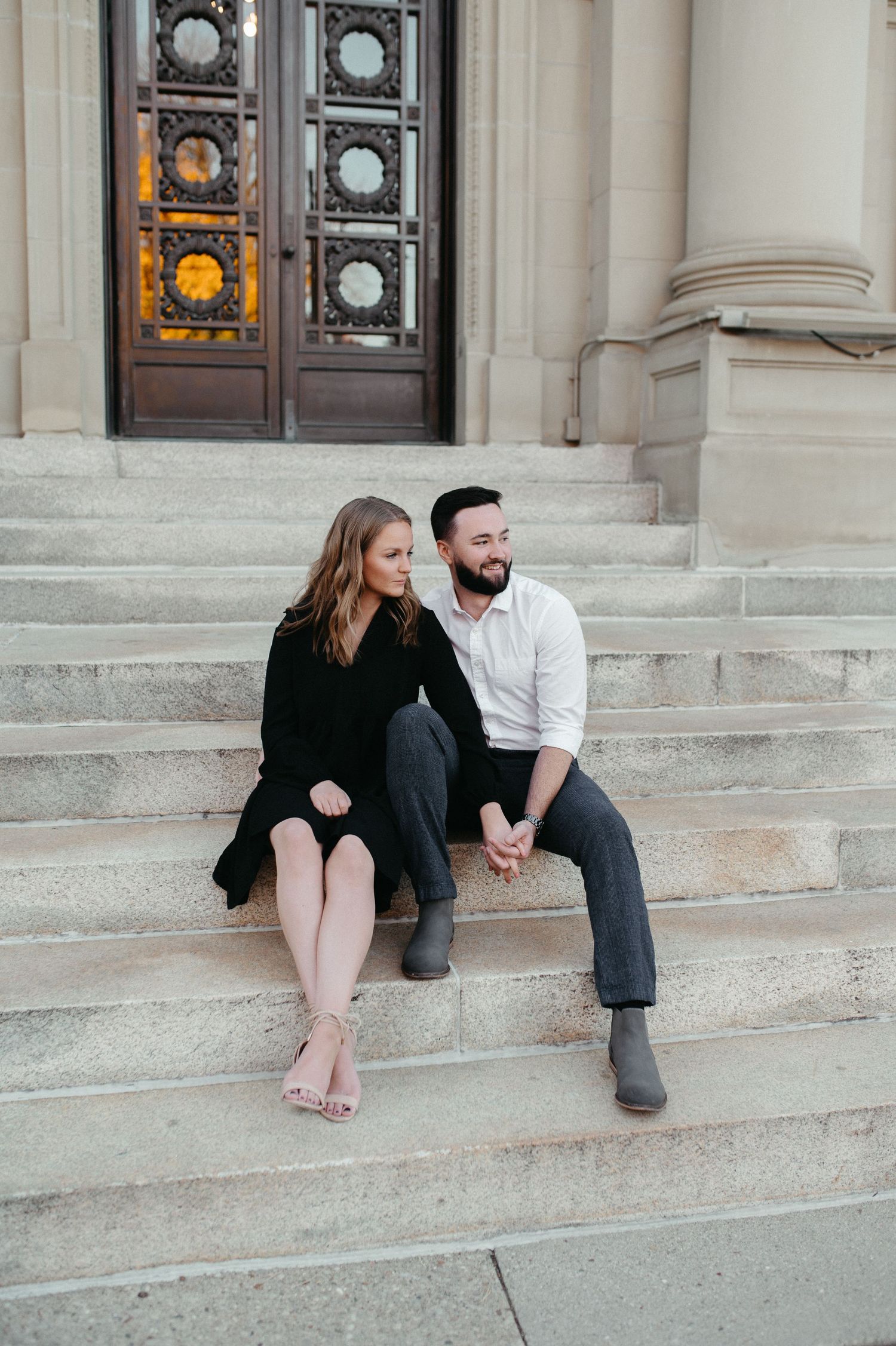 A couple sits casually on stone steps in front of decorative doors during an outdoor photoshoot.