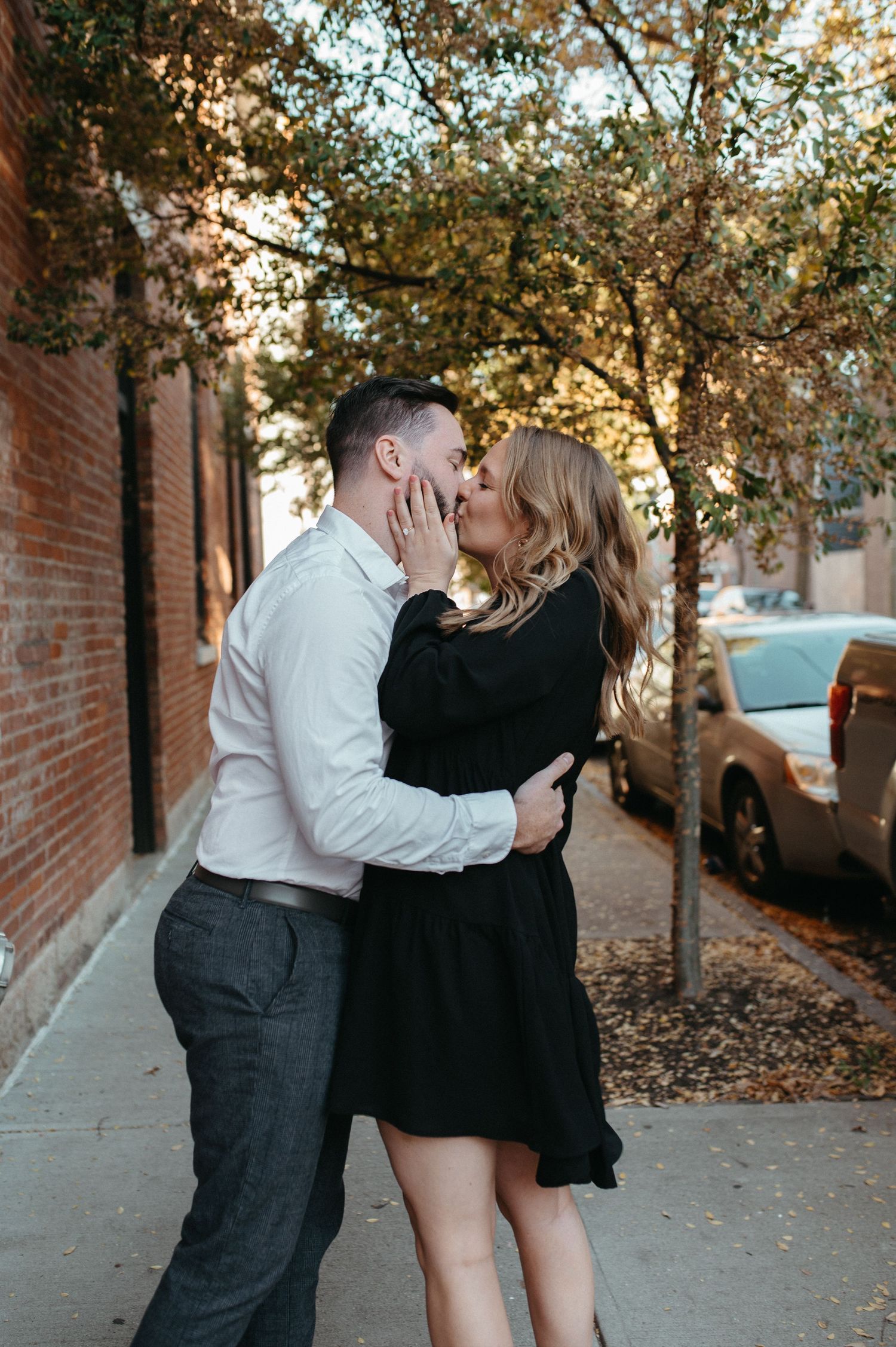 A couple in elegant attire embracing on an urban sidewalk with fall foliage overhead.