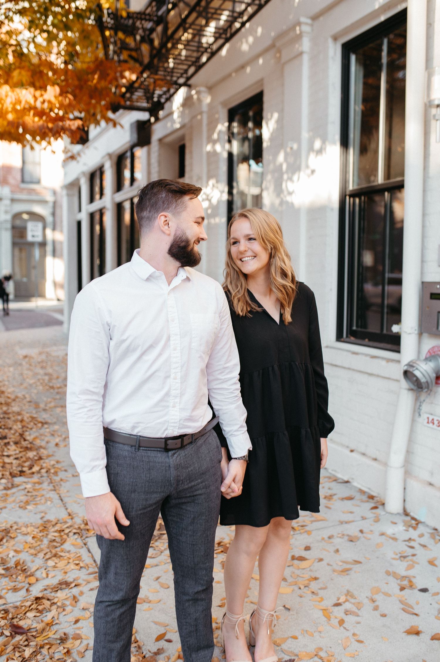 Couple holding hands while walking on autumn sidewalk with fallen leaves.