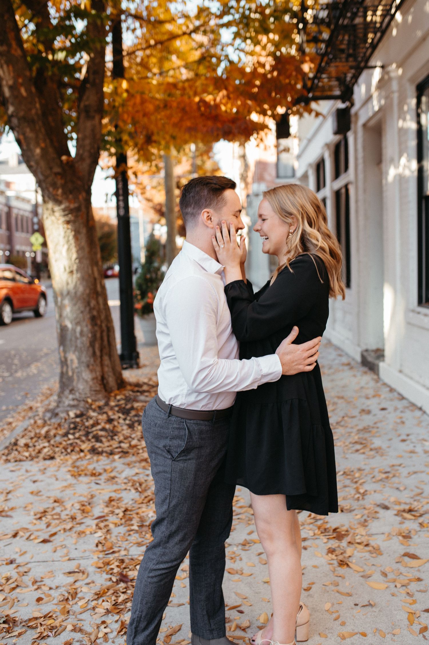Couple embracing on a leaf-covered sidewalk with autumn trees and urban buildings.