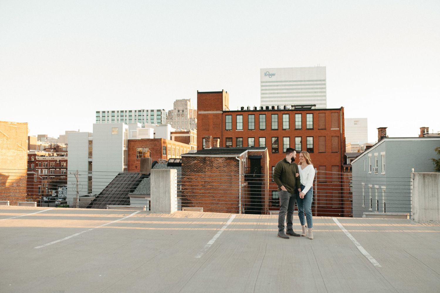 A couple stands together on a rooftop parking area with brick buildings in the cityscape.