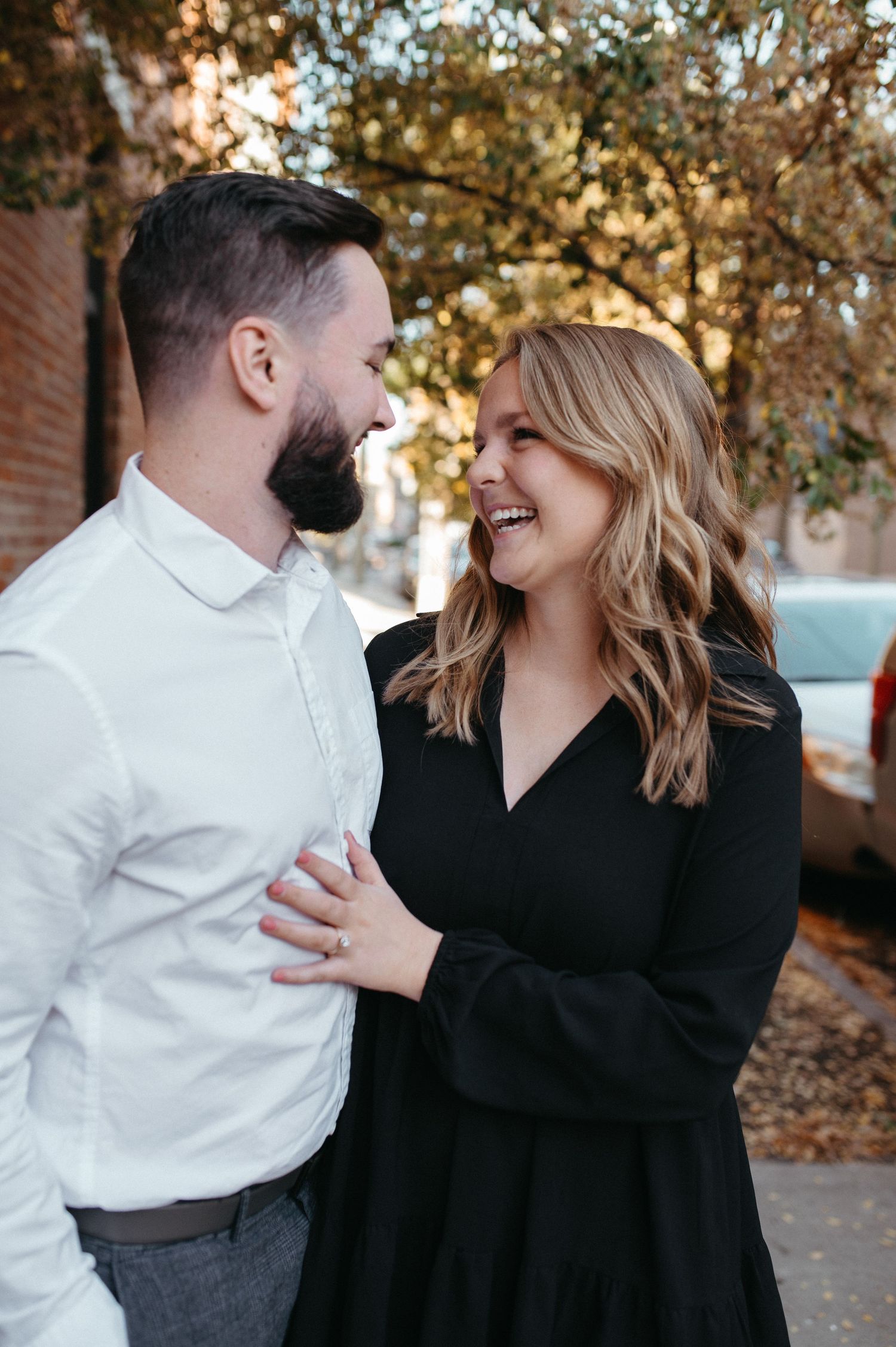 A candid moment between a couple laughing together against an urban backdrop with fall foliage.