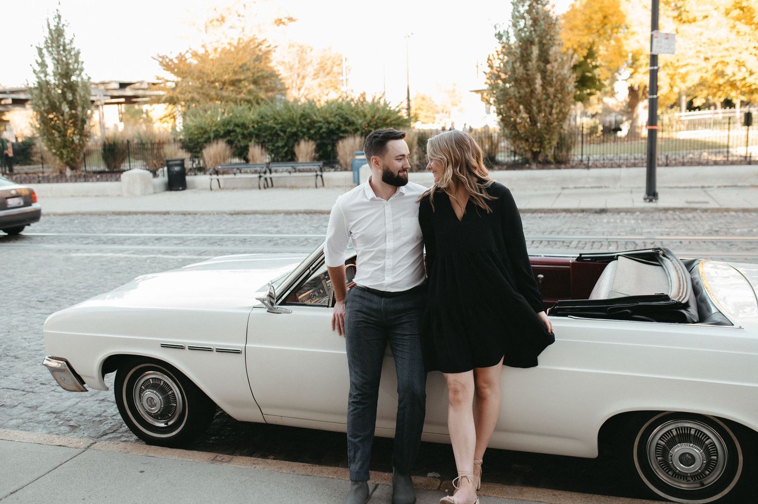 A couple poses next to a classic white convertible car on a street with autumn trees in the background.