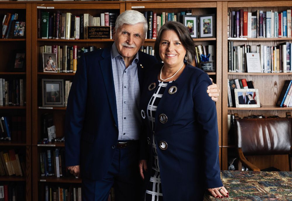 A couple poses together in front of a bookshelf-lined study in their home.