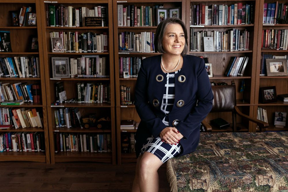 Professional portrait of person seated in navy blazer against library backdrop.
