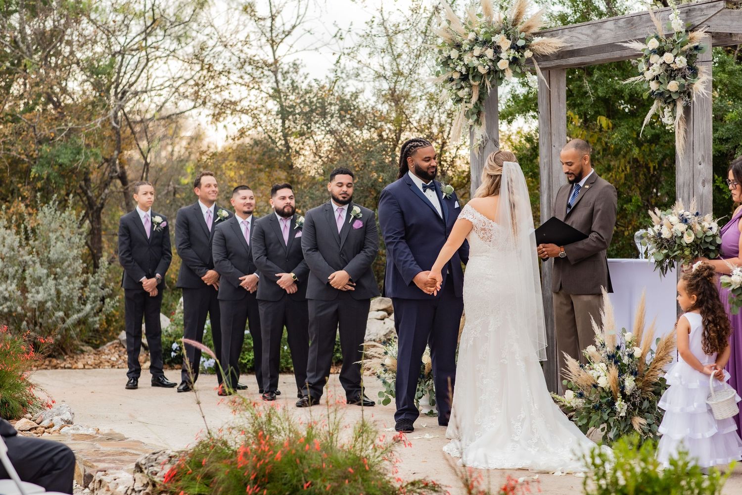 Outdoor wedding ceremony with bridal party in navy suits and elegant floral arch decor.