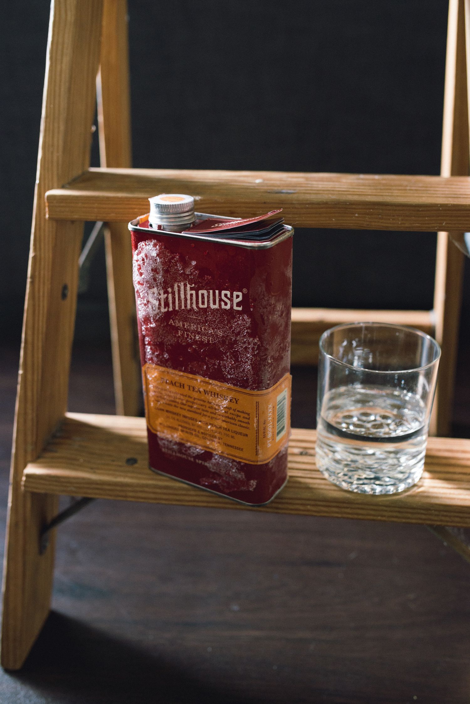 Wooden ladder with glass of water and red Stillhouse whiskey container against dark background.