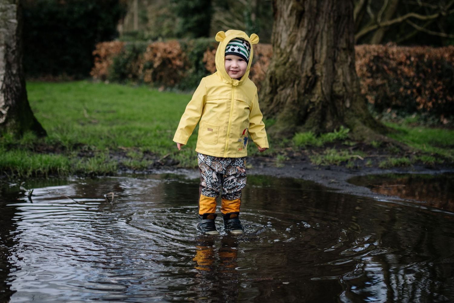 Child in yellow raincoat splashing in muddy puddle surrounded by trees.
