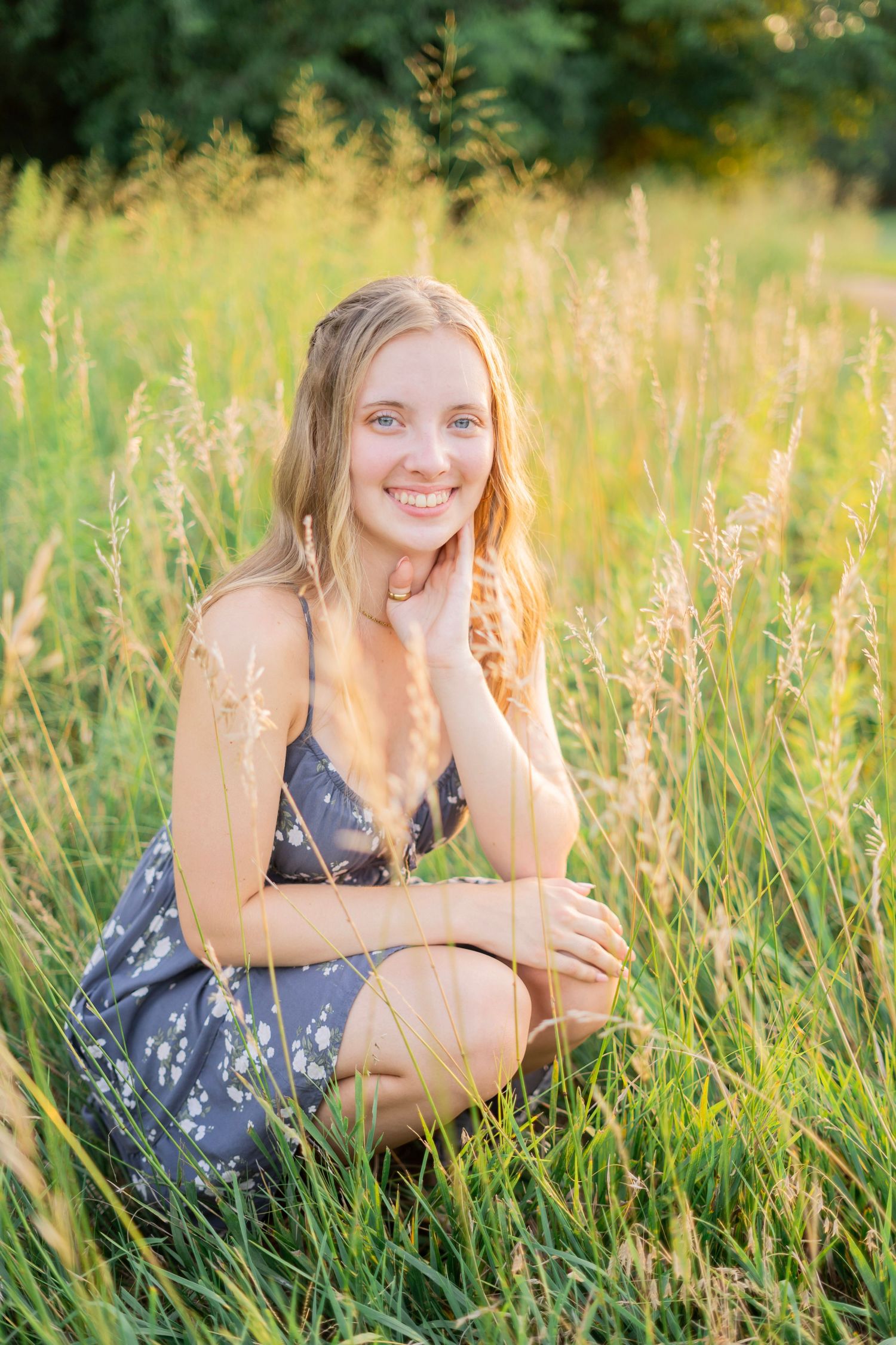 Smiling blonde teen girl in floral dress poses in golden sunlit tall grass field.