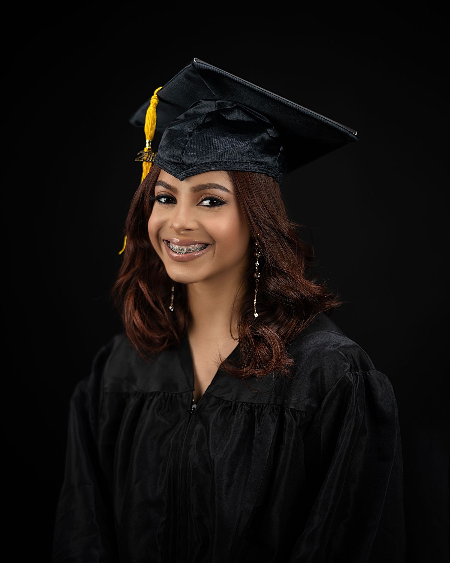 Graduate wearing black cap and gown with yellow tassel smiles against dark backdrop.