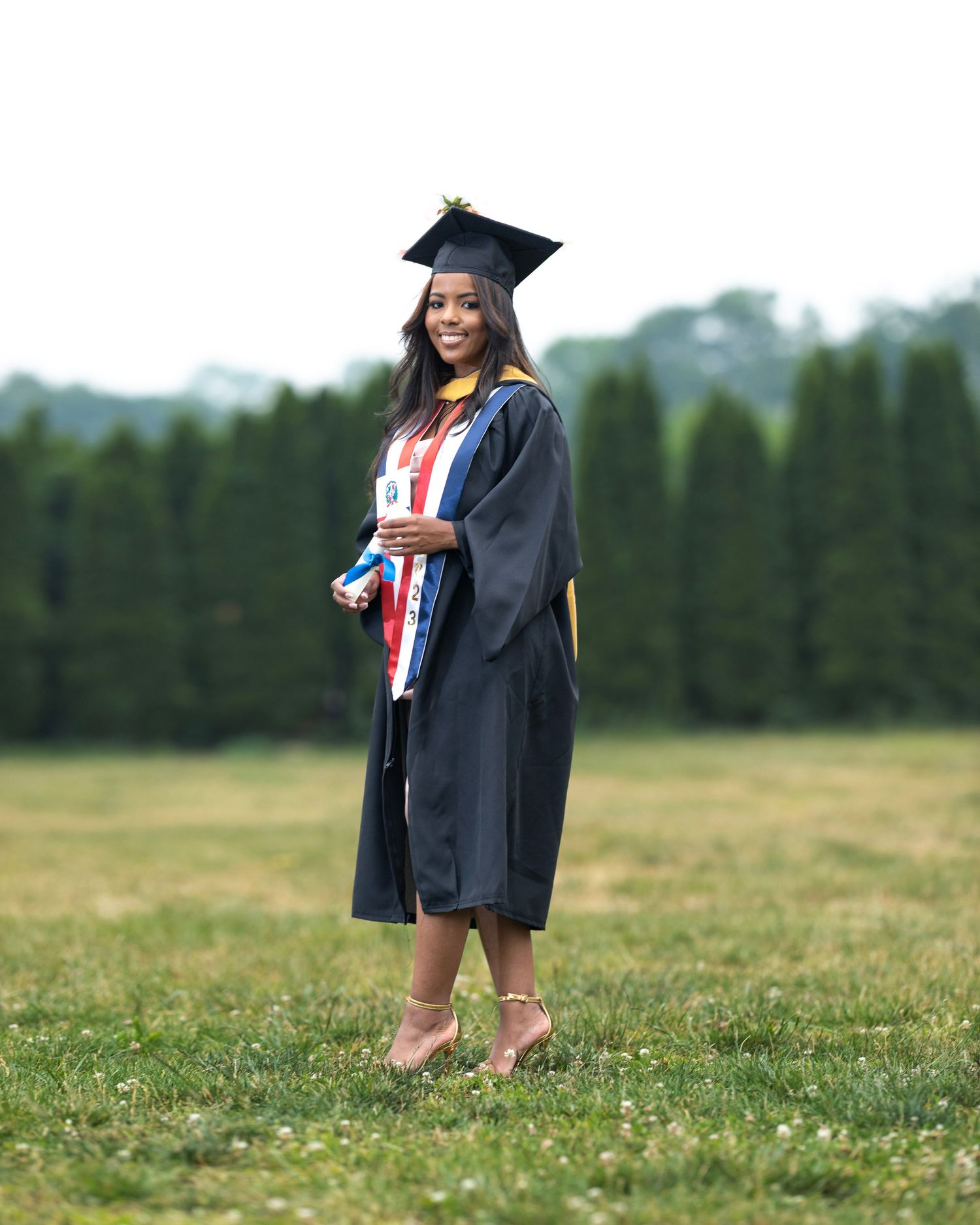 Graduate in academic regalia stands in grassy field with evergreen trees in background.