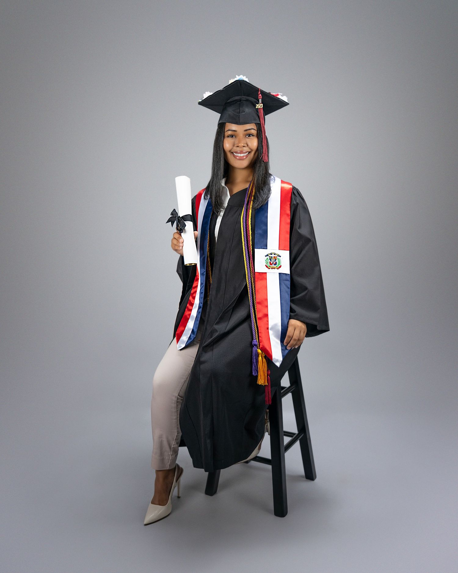 Graduate in black cap and gown with red stole poses seated on stool.
