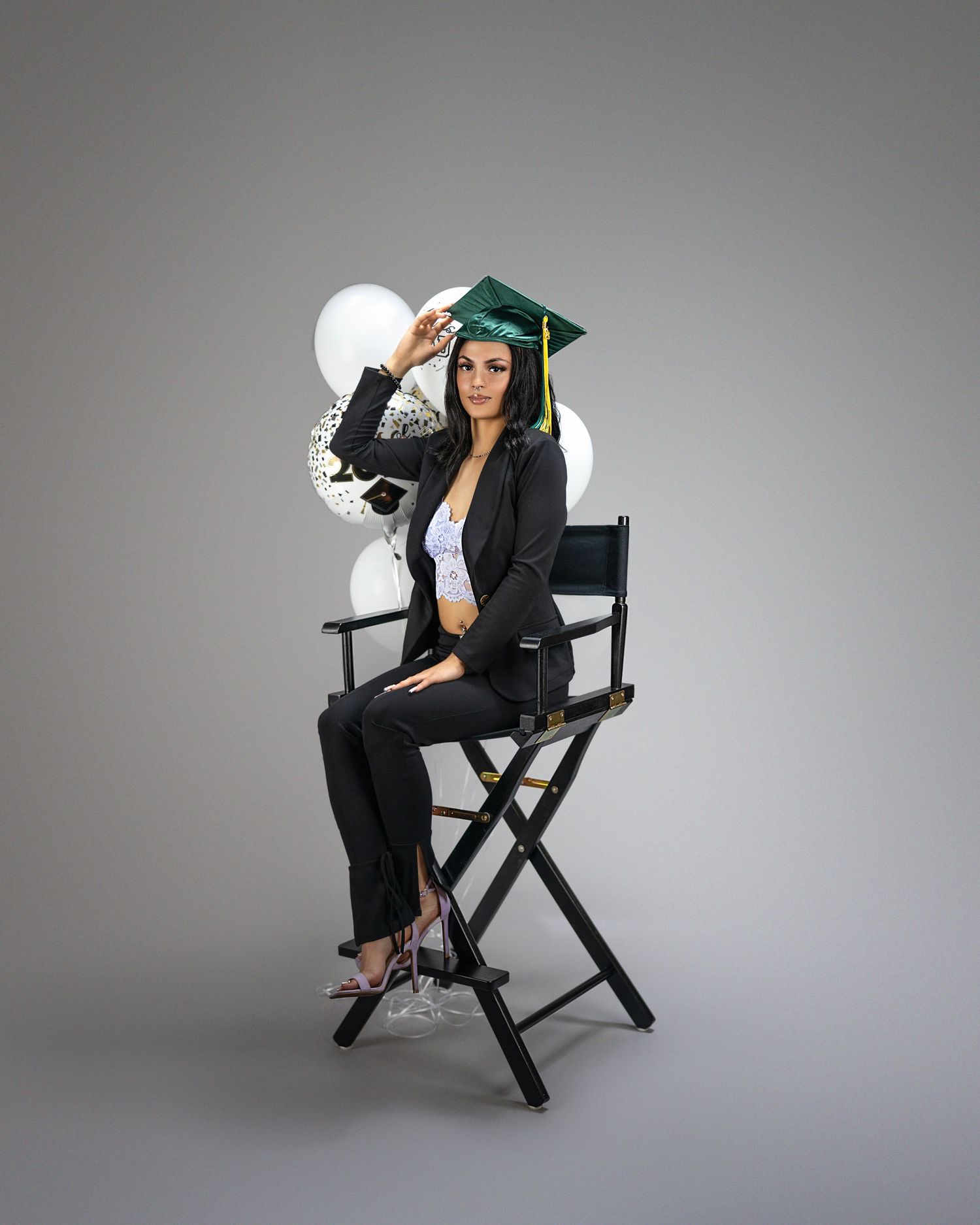 Graduate in cap and blazer sits on director's chair with white balloons in background.