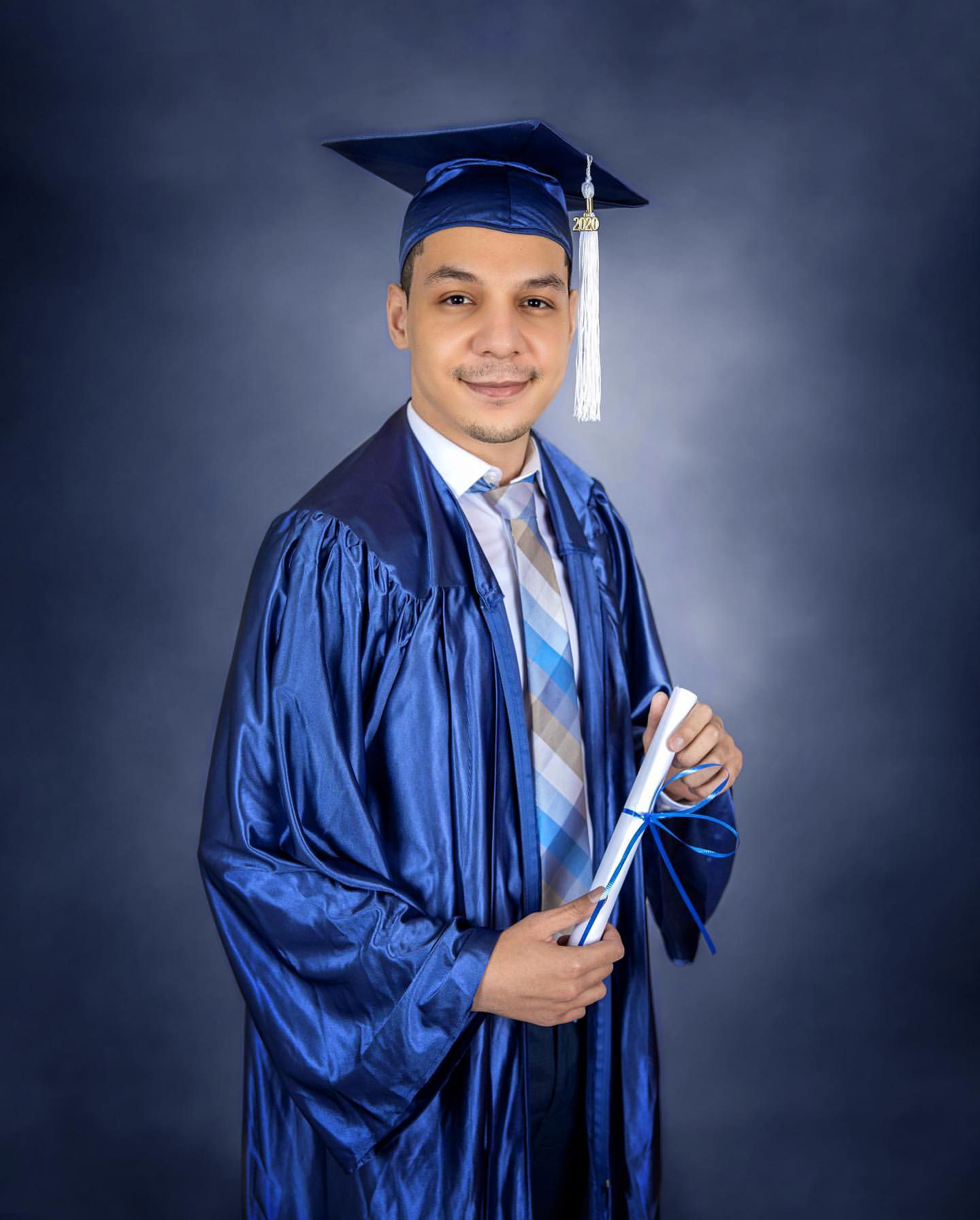 Graduate in royal blue cap and gown holding diploma poses against dark background.