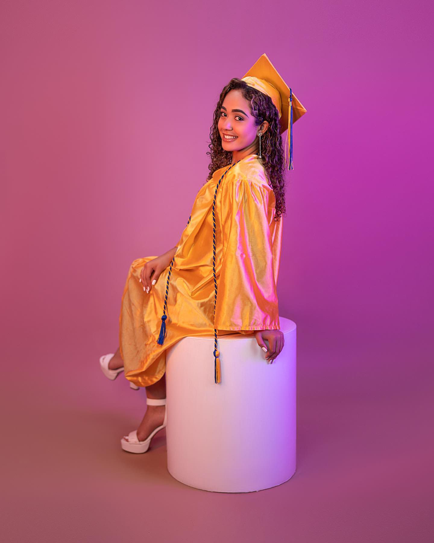 Graduate in yellow cap and gown poses on white pedestal against pink gradient background.