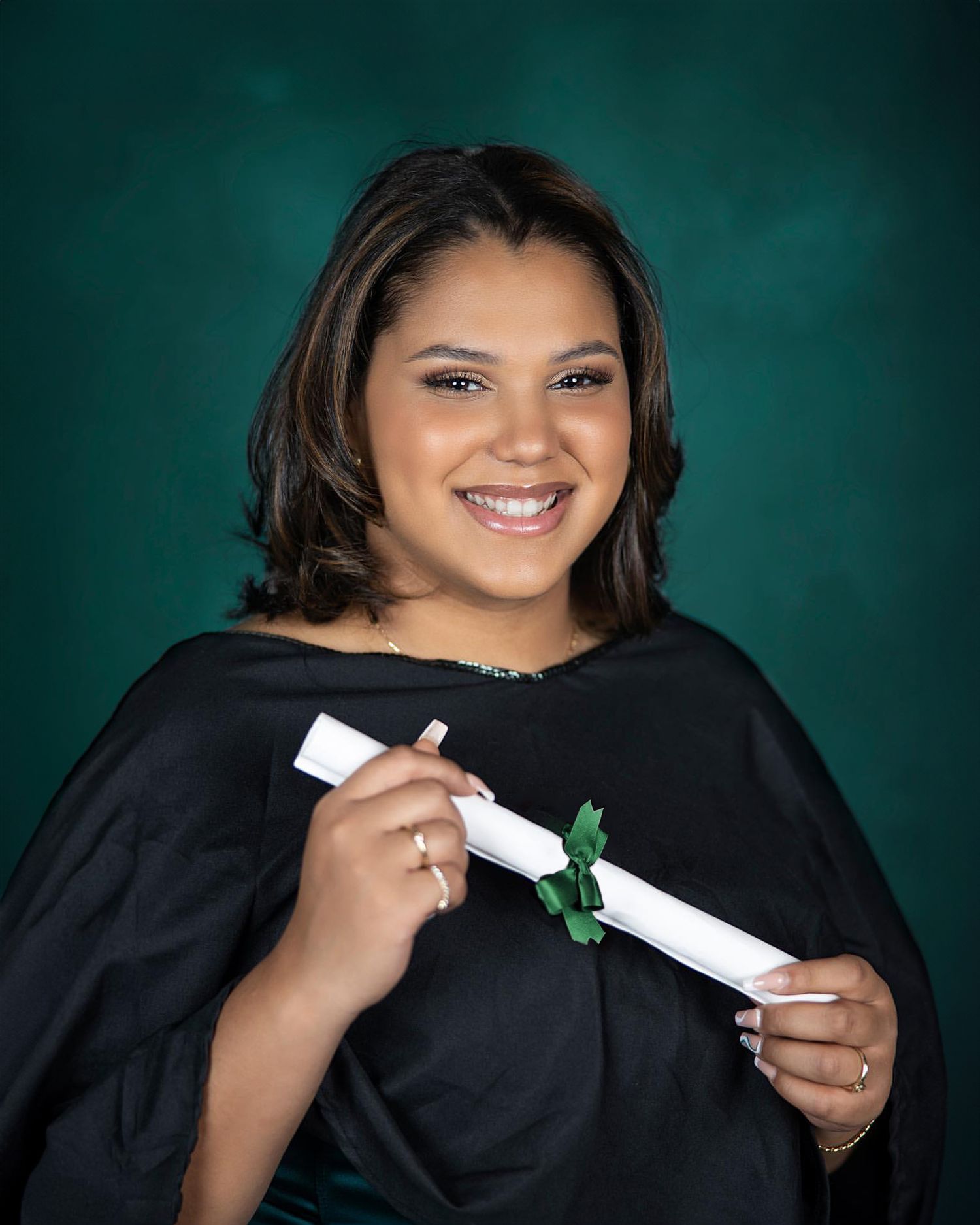 Graduate in black academic regalia holds diploma while smiling against green background.