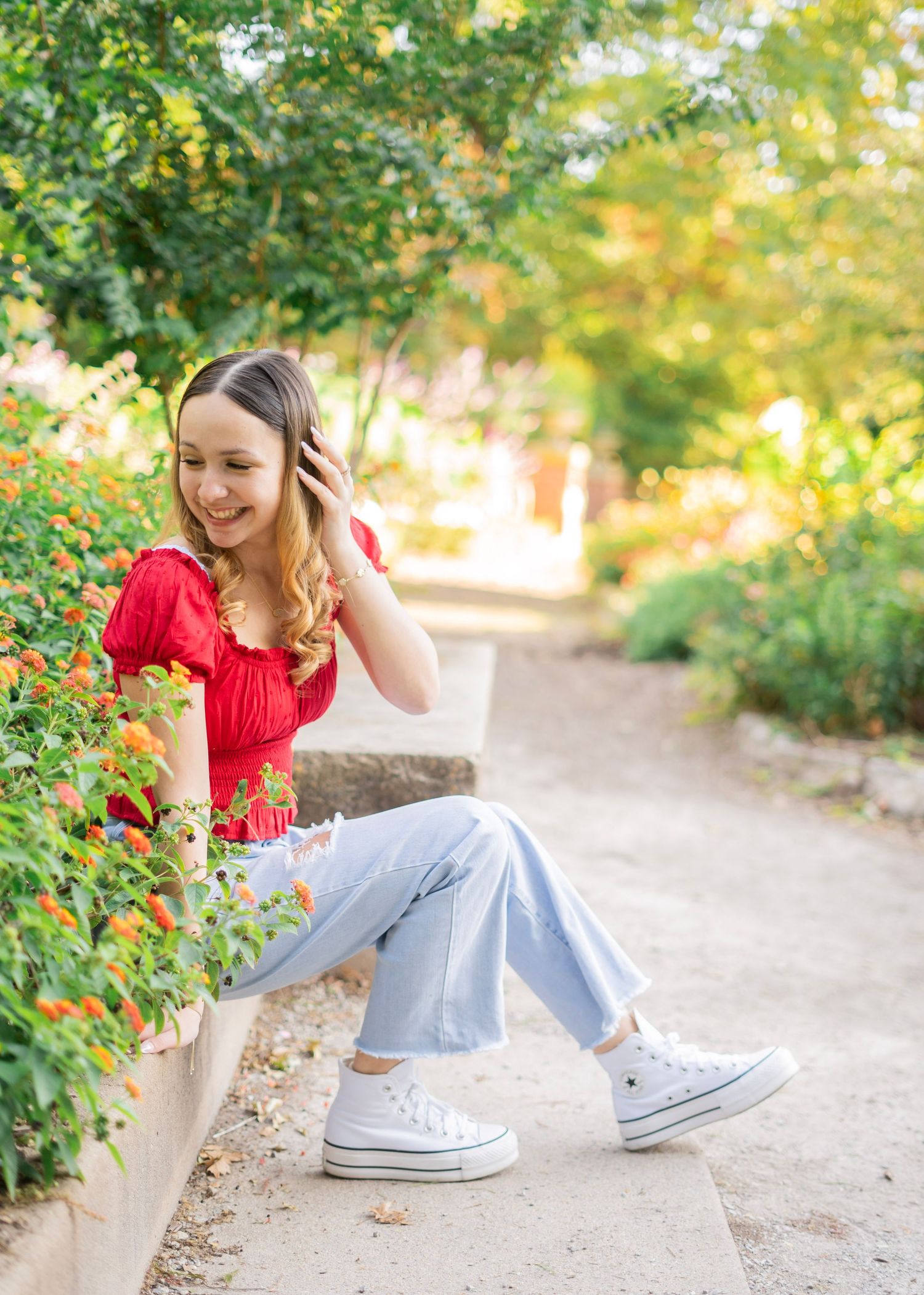 A smiling teenage girl in a red top and light jeans sits near orange flowers in a sunny garden.