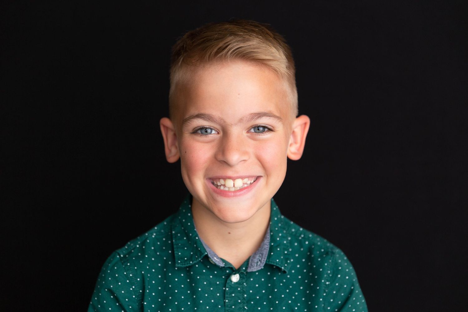 Portrait of young person in green polka dot shirt against black background with bright smile.