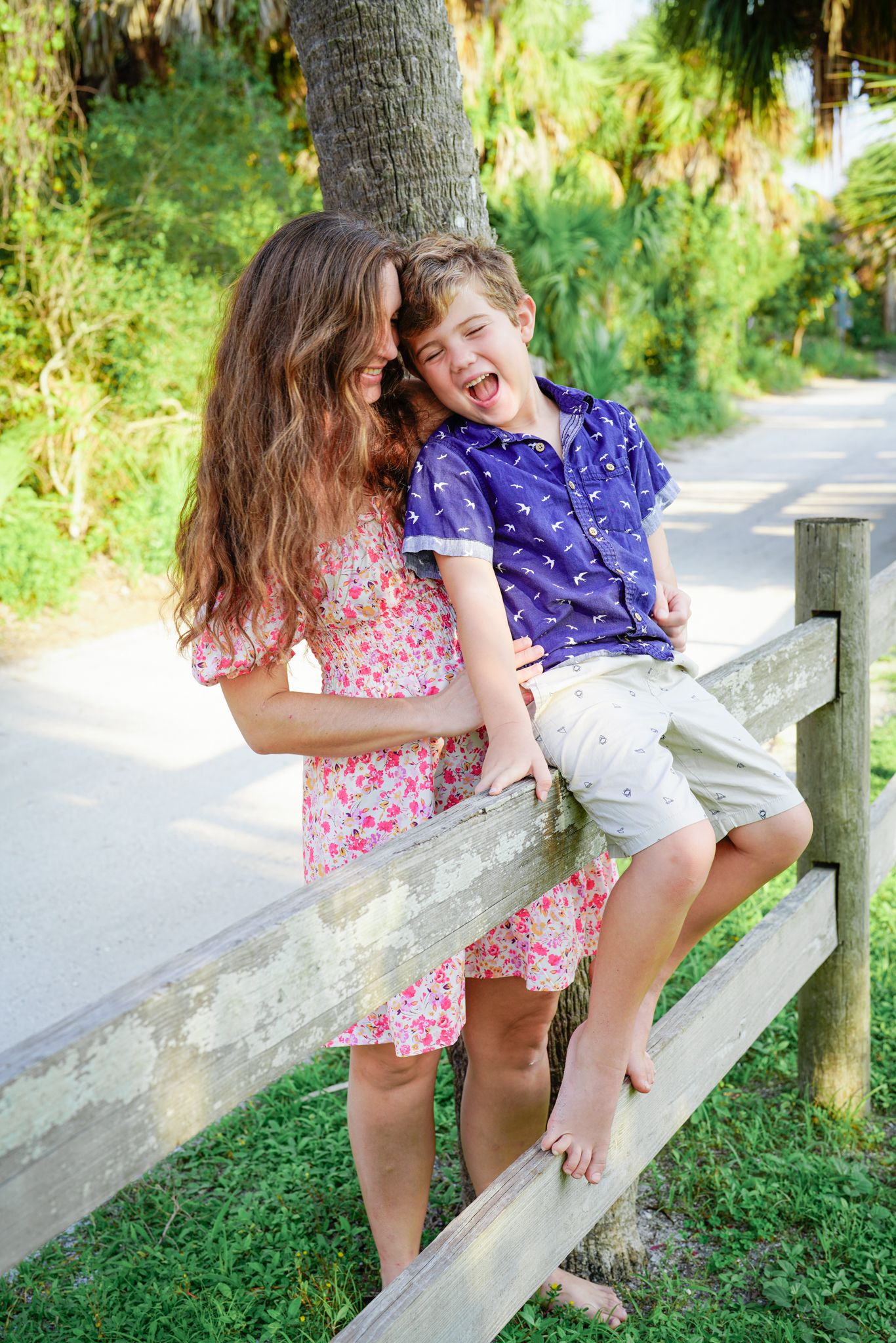 Young mother with long wavy hair wearing a floral dress giggling with her young son sitting on fencepost at beach area.
