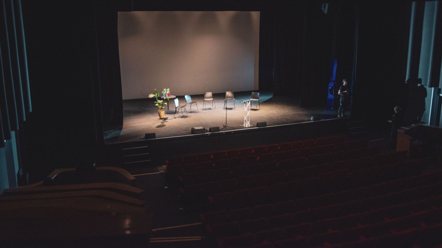 Small group of performers on a spotlit stage in an empty red-seated theater auditorium.