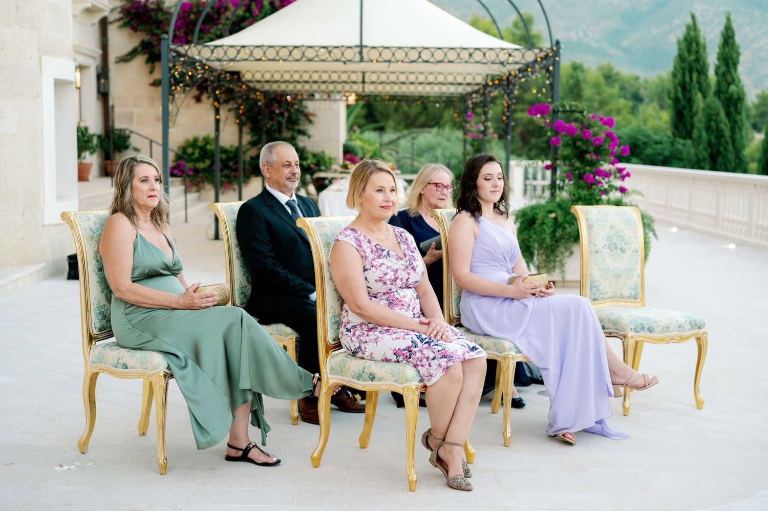 Wedding guests seated on gold chairs at outdoor Mediterranean venue.