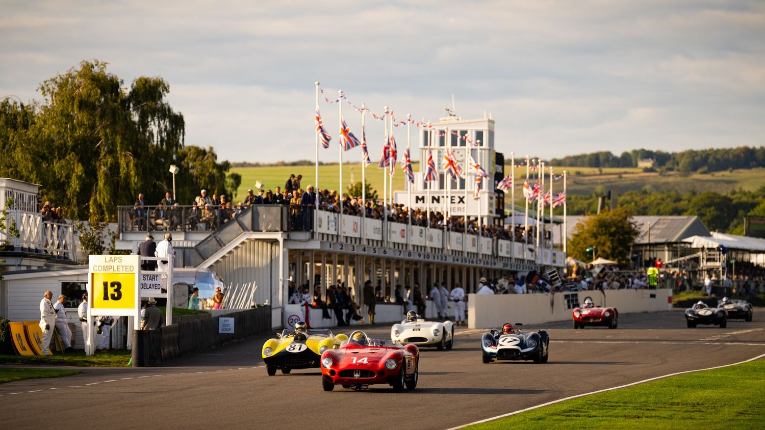 Vintage race cars line up at Goodwood Revival with classic buildings and flags in background.