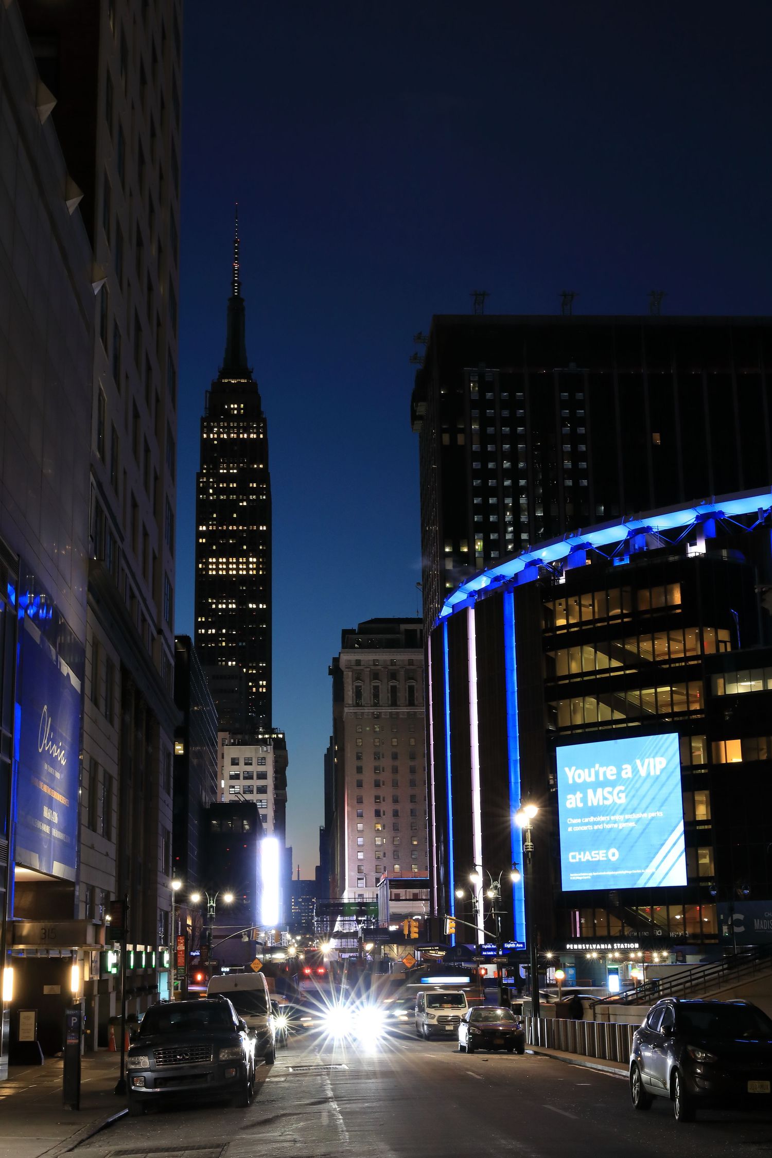 Empire State Building lit up at night with blue illuminated buildings and traffic on NYC street.
