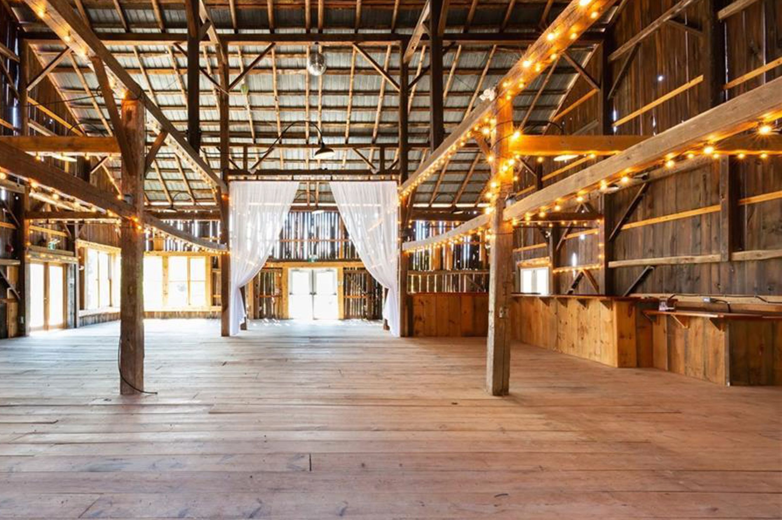 Interior of a rustic barn venue with wooden beams, string lights and white fabric draping.