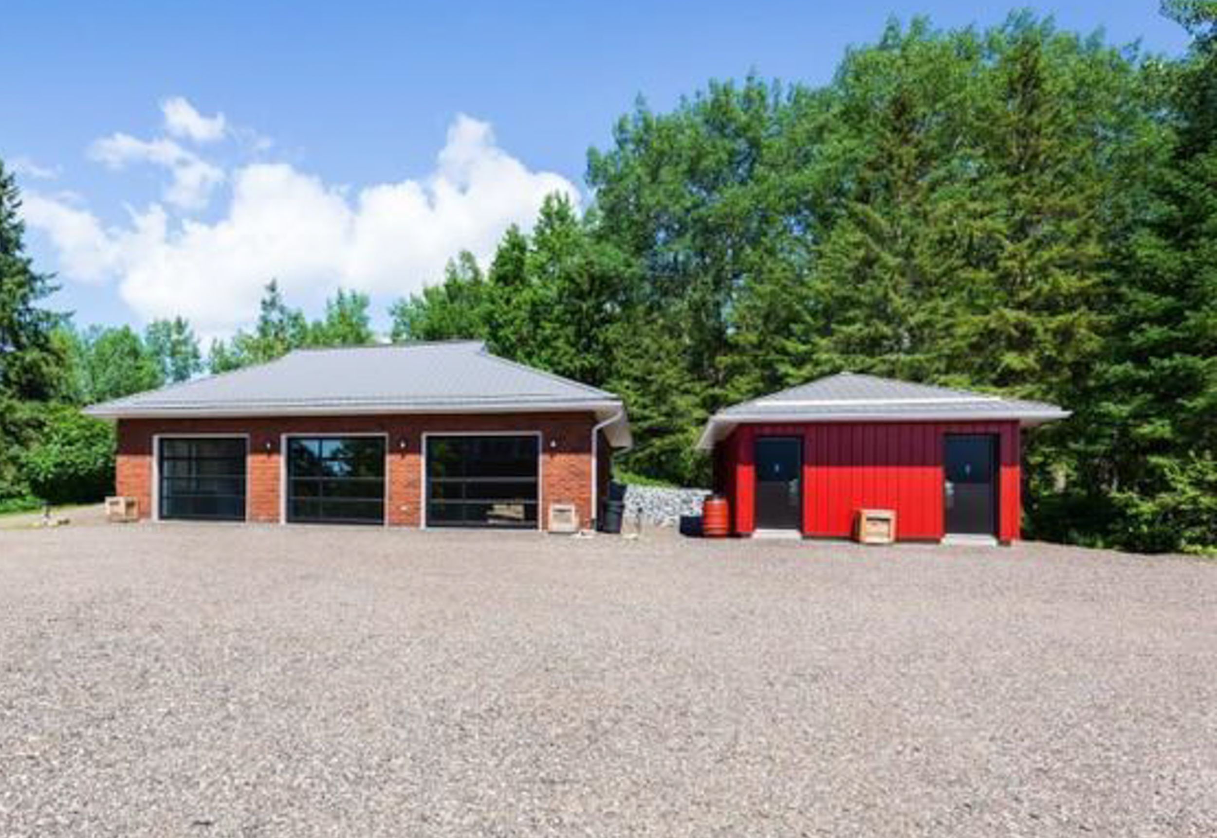 Red brick garage building with multiple doors next to a smaller red outbuilding on gravel lot.