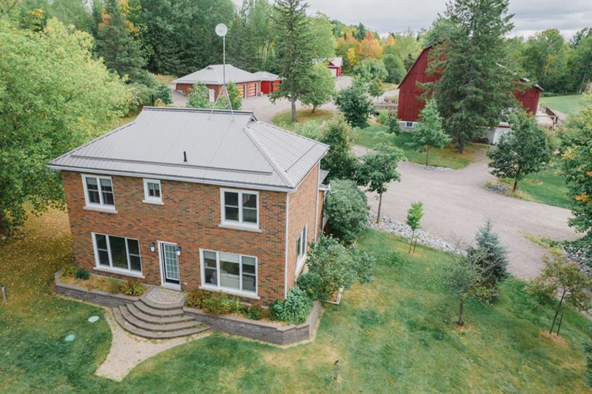 Two-story brick house with gray roof surrounded by trees and landscaping in a rural setting.