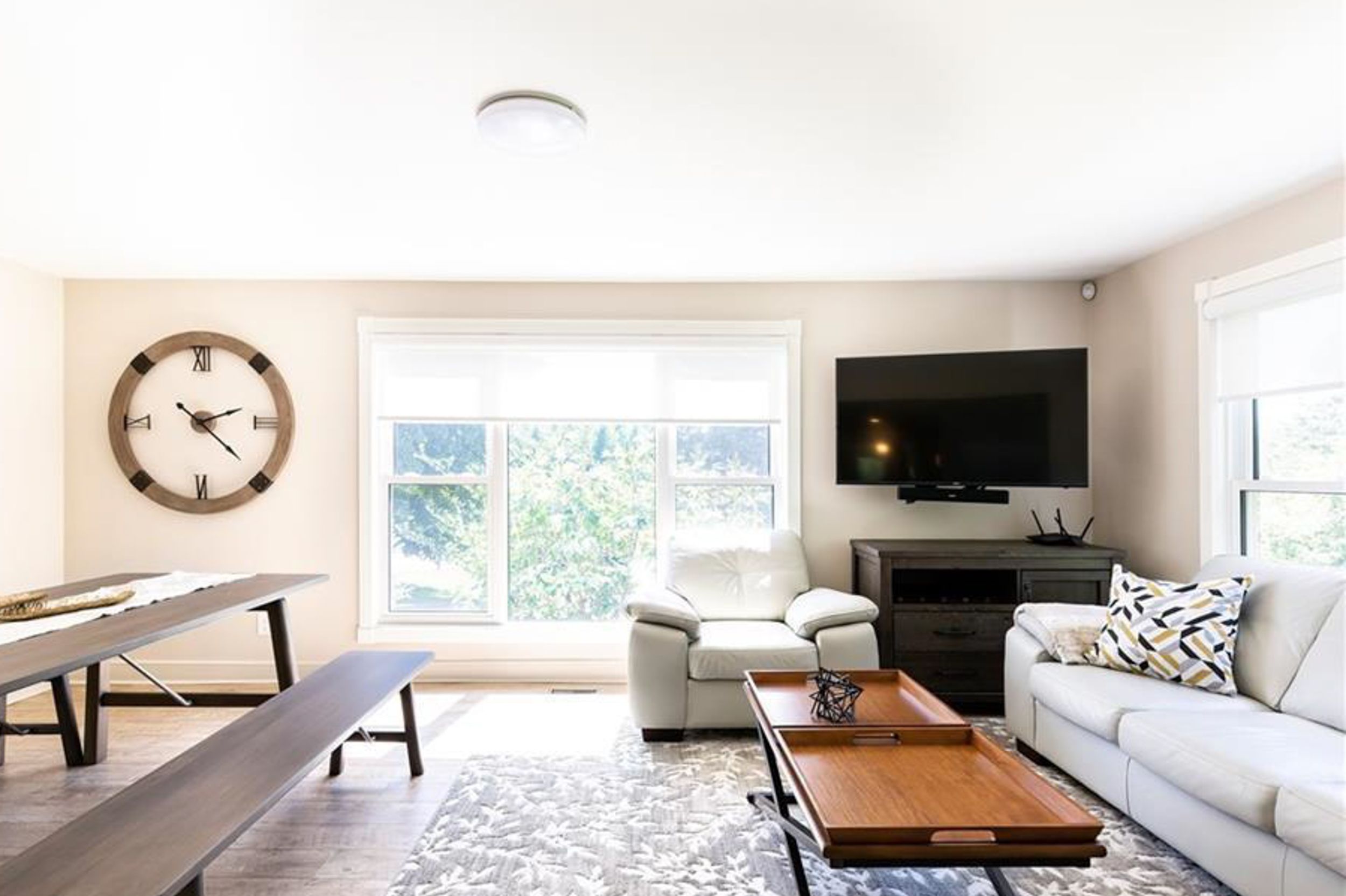 Modern living room featuring white couch, wall-mounted TV, oversized clock, and large windows.