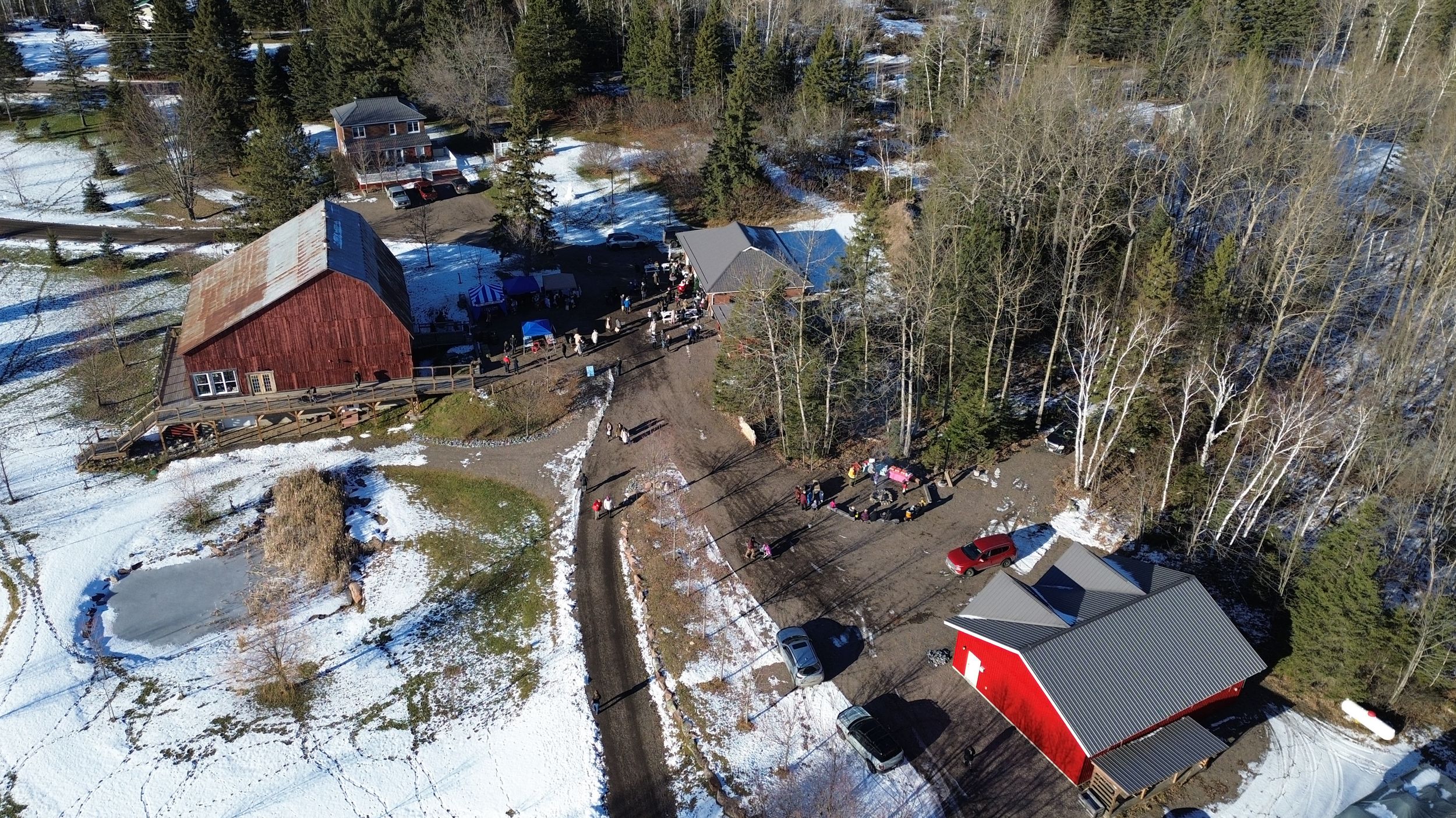 Aerial view of winter gathering at rustic red barn with parked cars and snowy grounds.