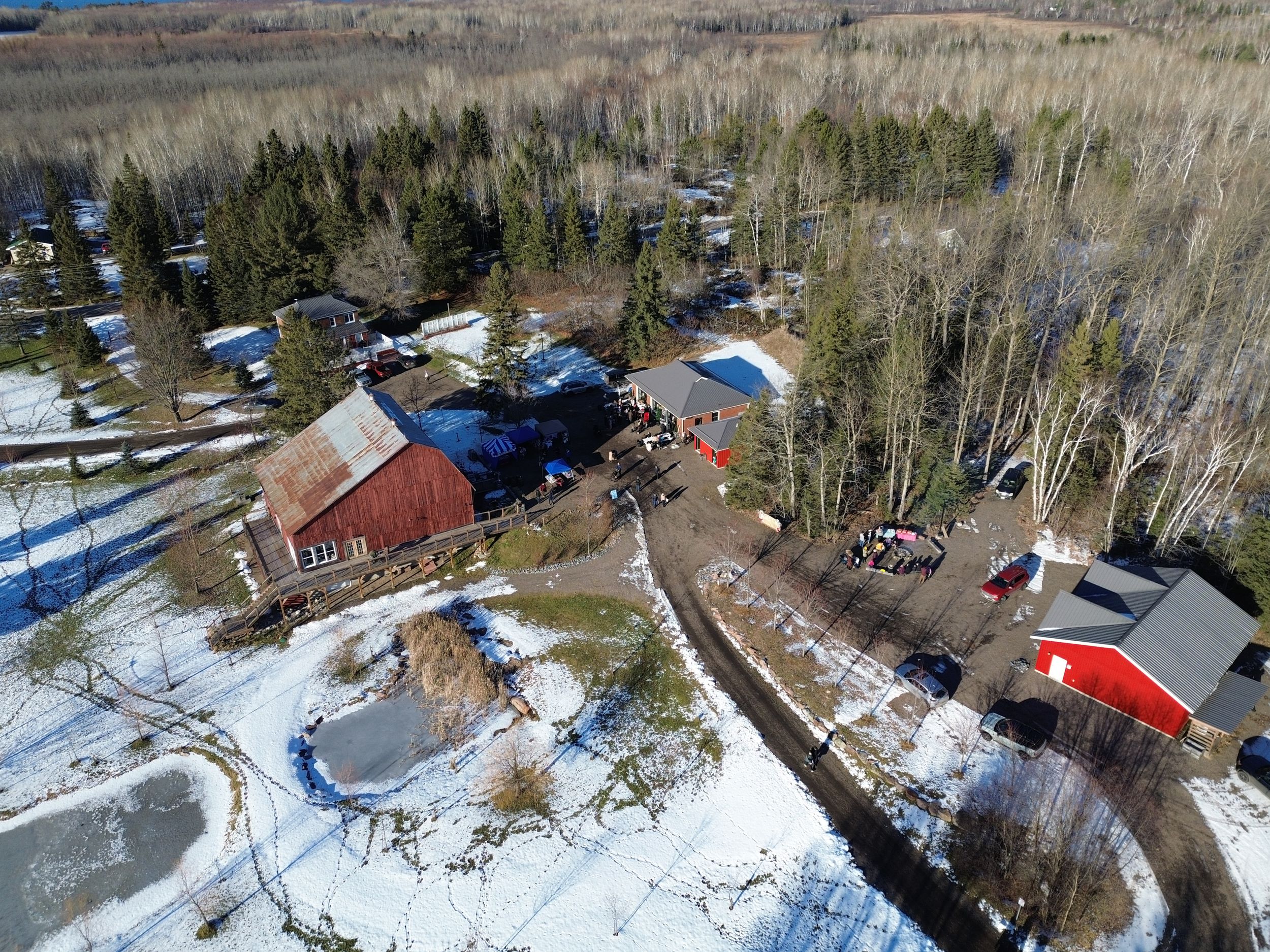 Winter scene of rural property with red barn, outdoor rink, and parking area surrounded by woods.