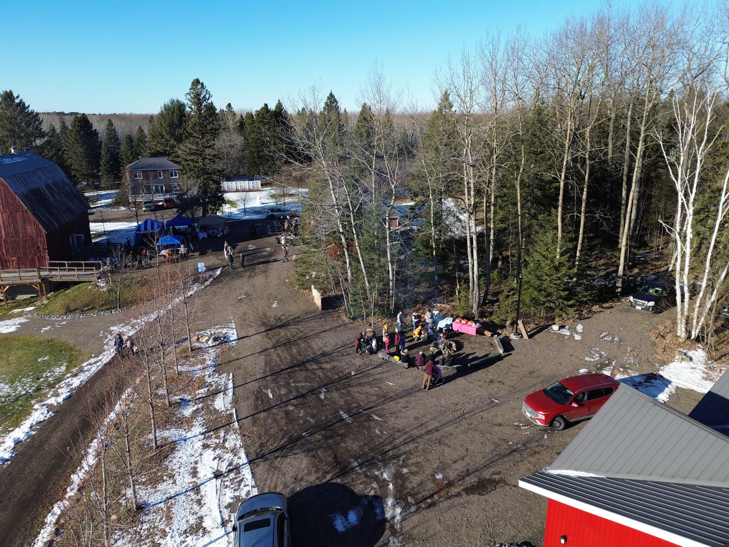 Outdoor winter gathering area near barn buildings with parked vehicles and snow-lined paths.