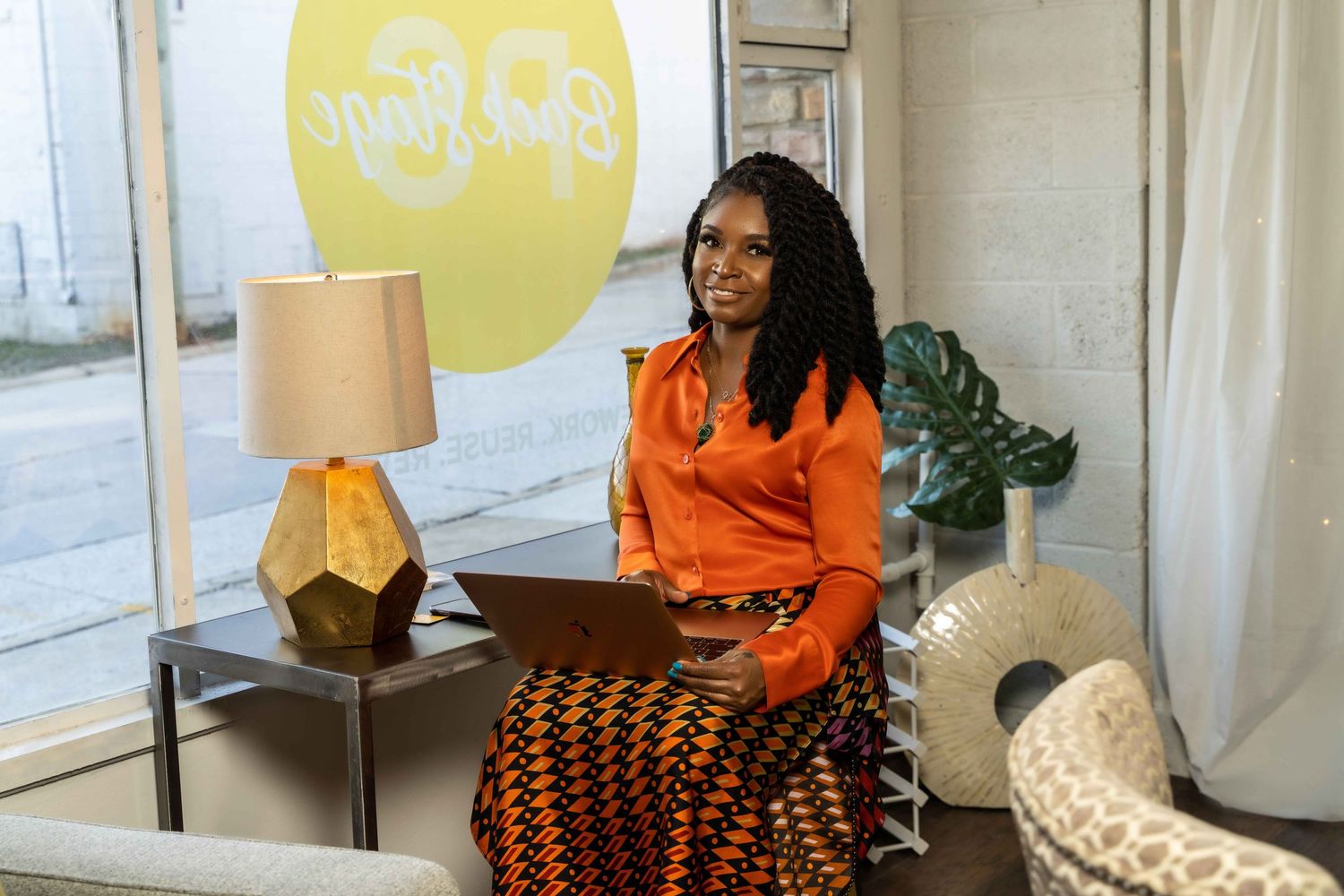 Person in orange top seated beside geometric lamp with yellow circular artwork in background.