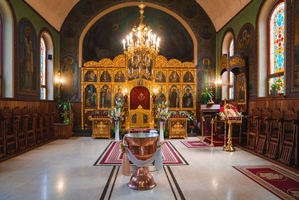 Interior of an ornate Orthodox church with chandelier, iconostasis, and red carpet leading to the baptismal font.