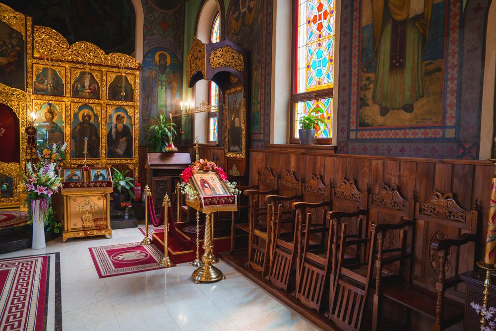 Wooden church pews beside an ornate golden iconostasis with stained glass windows and red carpeting.