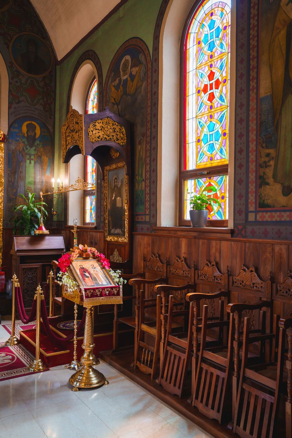 Stained glass windows illuminate wooden chairs and religious decor in an Orthodox church interior.