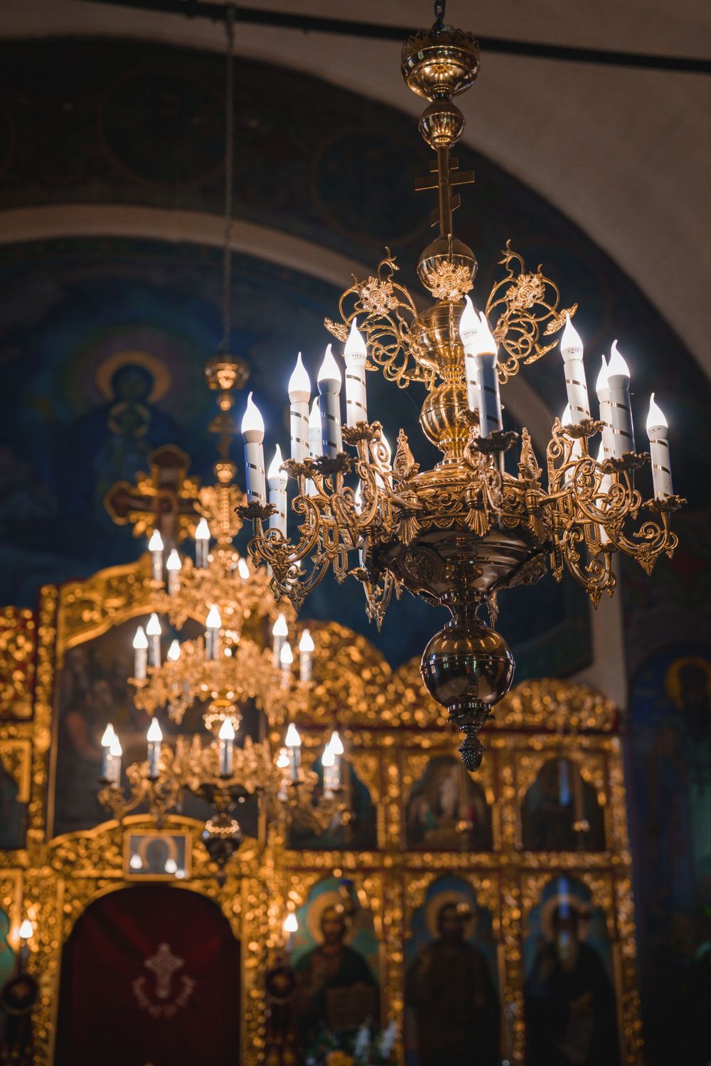 Elegant brass chandelier with crystal details hanging in front of a golden Orthodox church iconostasis.