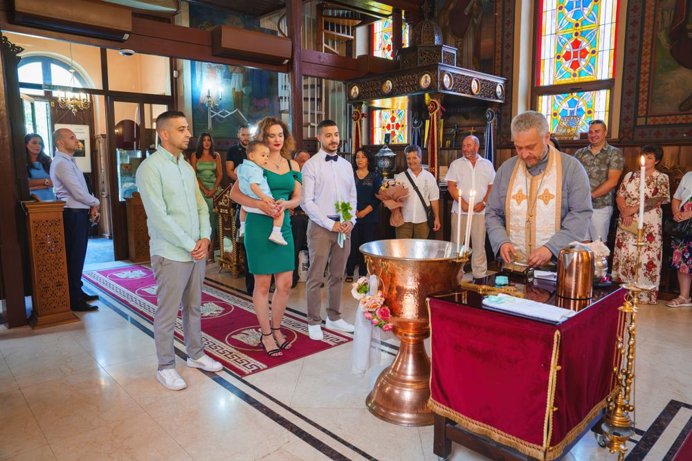 An Orthodox Christian baptism ceremony taking place inside a church with stained glass windows.