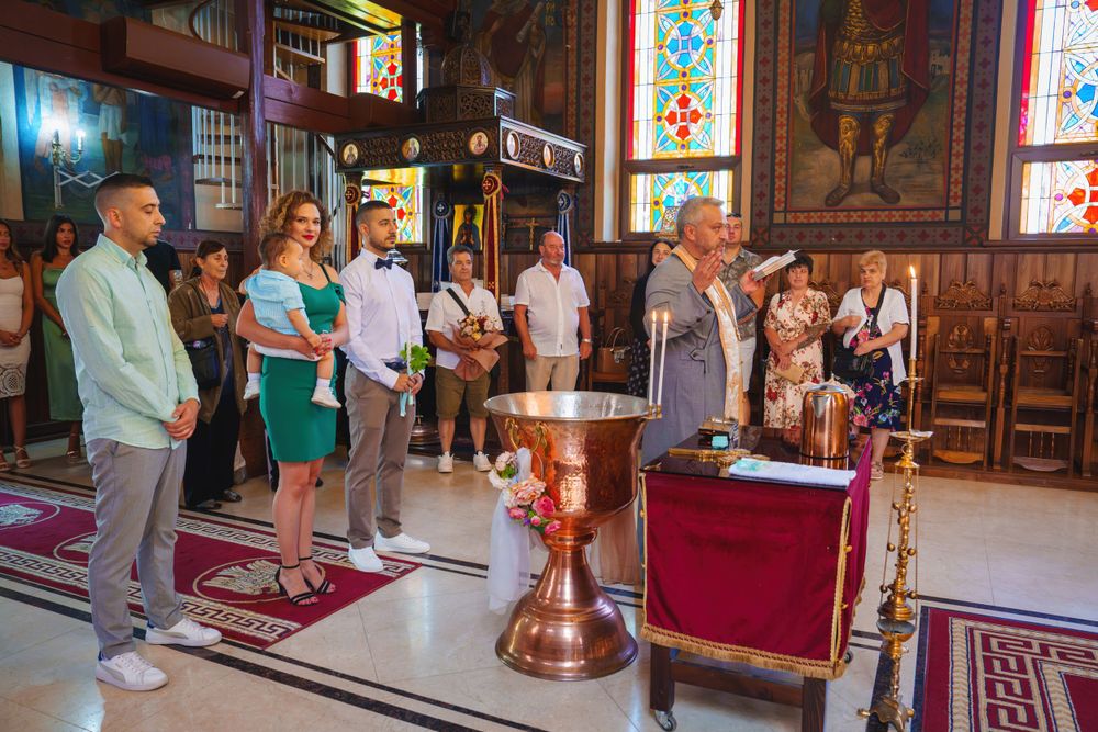 Religious ceremony participants gathered around a baptismal font in a traditional church setting.
