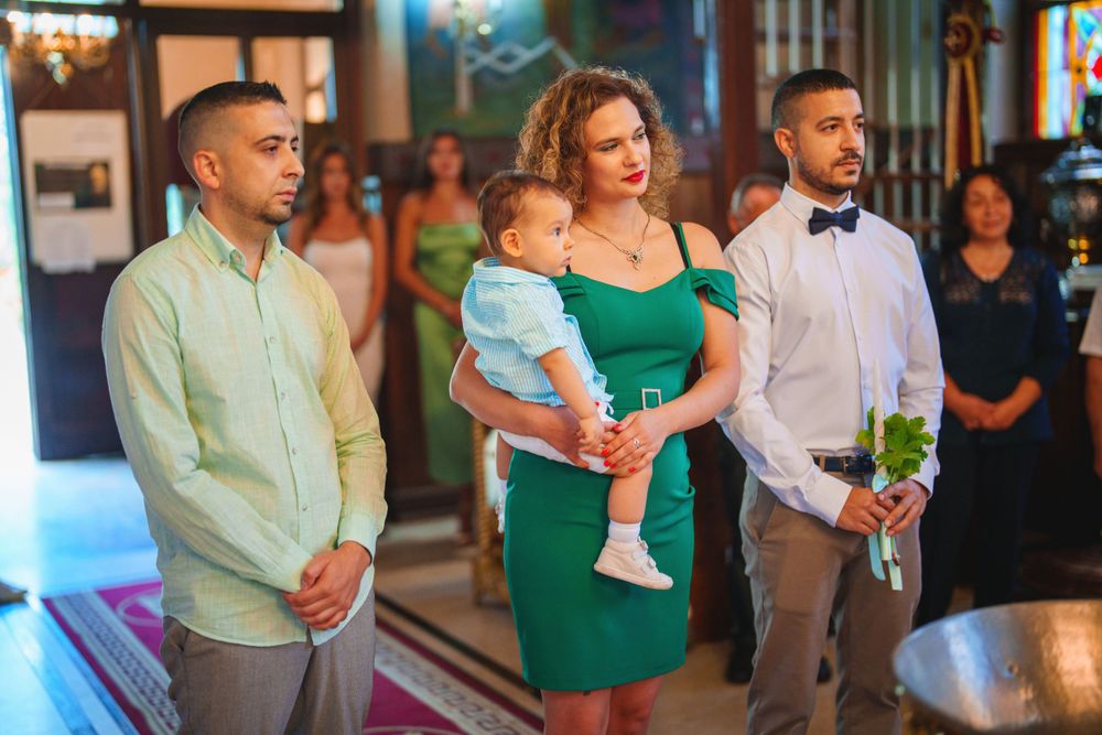 Baptism ceremony participants in formal attire standing in front of church stained glass windows.