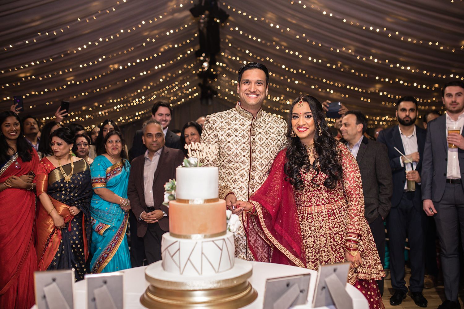 Wedding couple cuts their elegant tiered cake under string lights surrounded by guests in traditional attire.